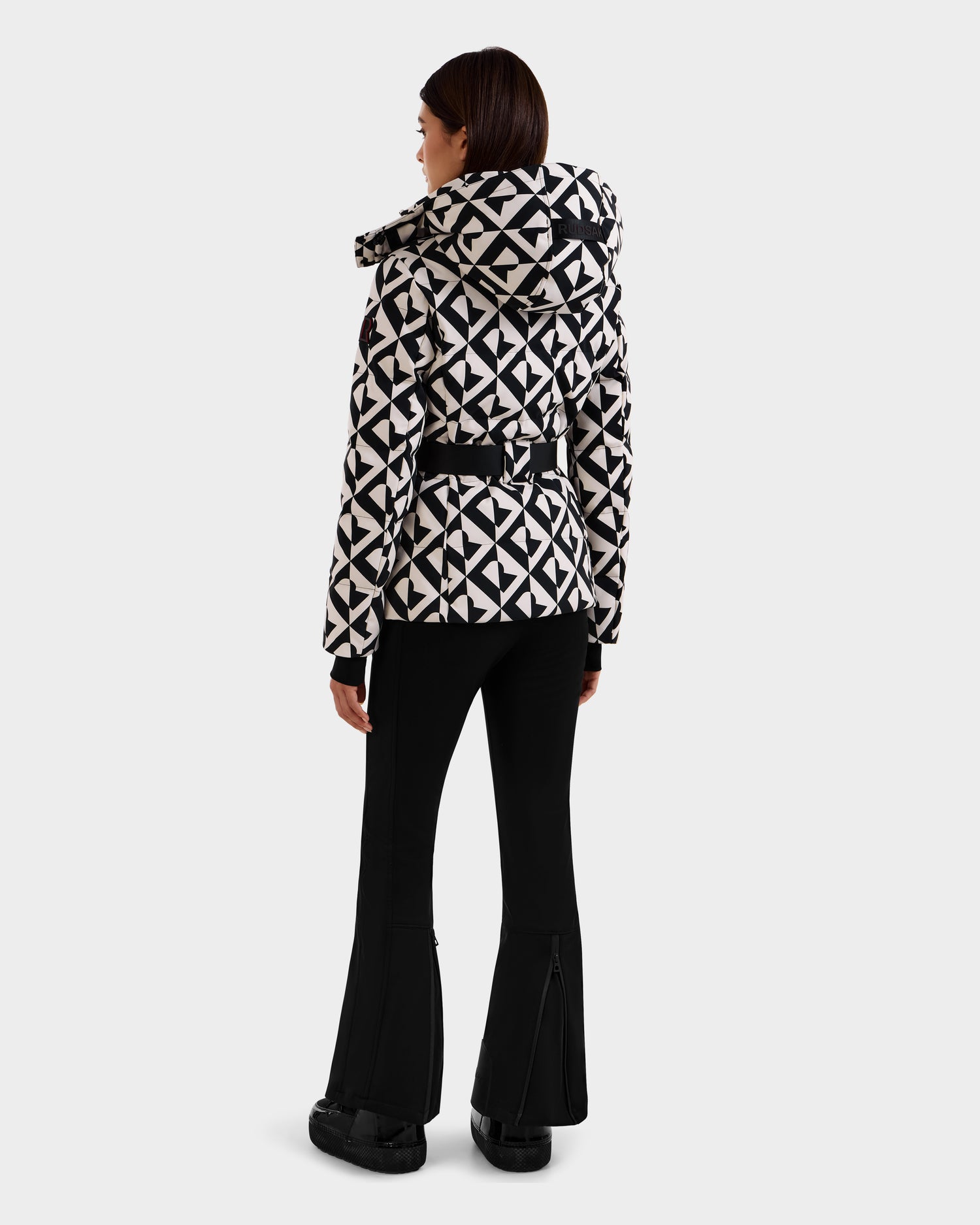 A woman faces away, wearing the Rudsak JOANNA P - 8125623 Vanilla Monogram women’s down ski jacket with hood, belt, and ski-pass pocket, paired with black flared pants and shoes against a plain background.