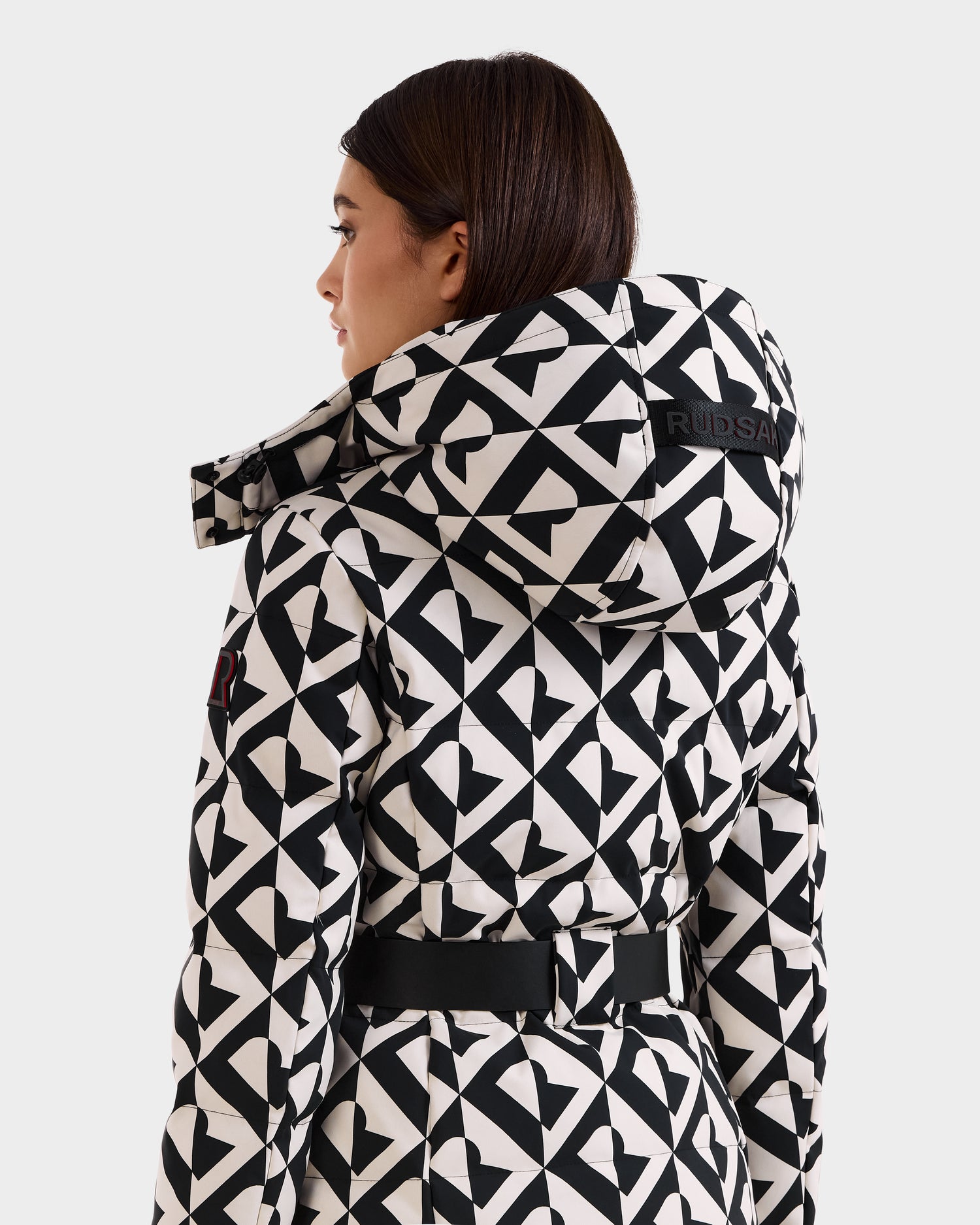 A woman with straight brown hair is shown from the back, wearing the Rudsak JOANNA P - 8125623 Vanilla Monogram coat. The hood features "RUDSAK" branding and a ski-pass pocket, with a belted black and white geometric pattern.
