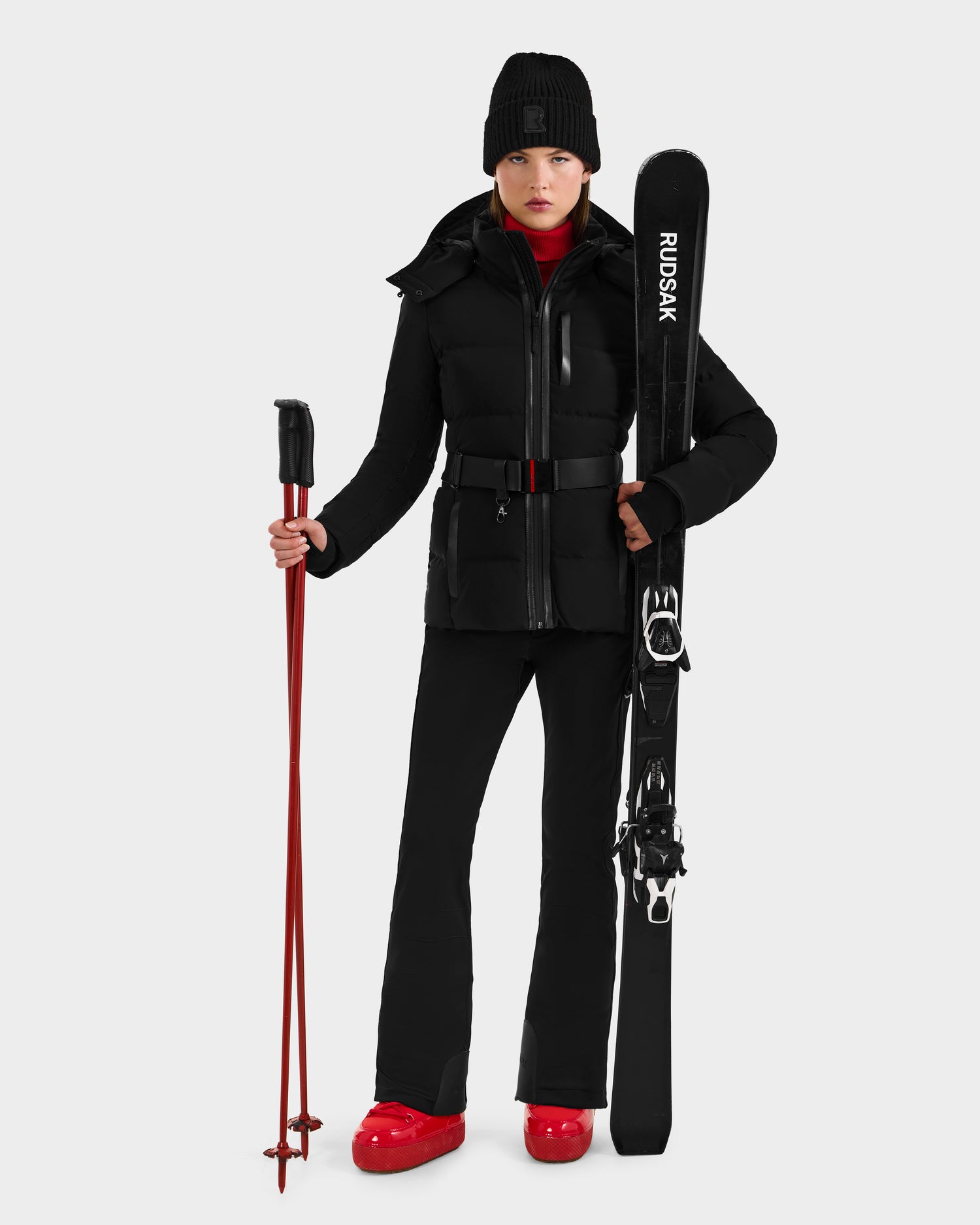 Wearing the JOANNA - 8124788 Black by Rudsak, a person holds black Rudsak skis and red poles, sporting red shoes against a plain background. This item is available for pre-order and ships by November 30th.