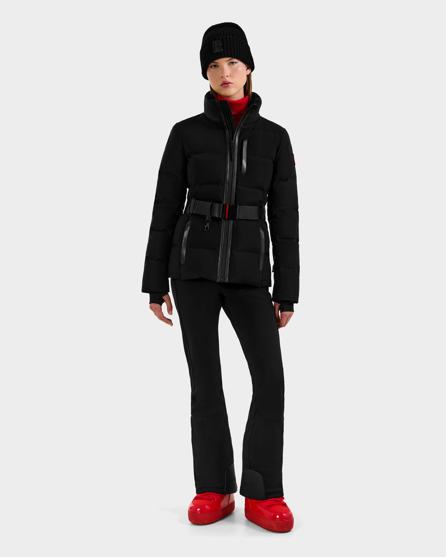 A woman wearing the Rudsak JOANNA - 8124788 Black women’s down ski jacket with a removable belt, paired with black pants and red boots, stands against a light gray background.