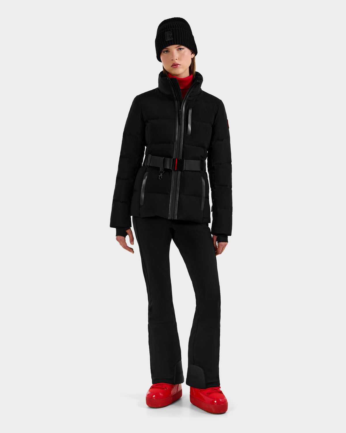 A woman wearing the Rudsak JOANNA - 8124788 Black women’s down ski jacket with a removable belt, paired with black pants and red boots, stands against a light gray background.
