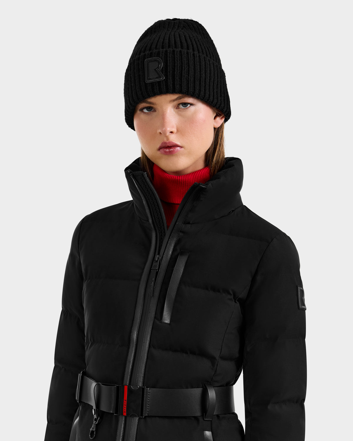 A woman wearing the Rudsak JOANNA - 8124788 Black women's down ski jacket with RECCO® rescue tech stands against a light background, paired with a red turtleneck sweater and black knit beanie, looking at the camera.