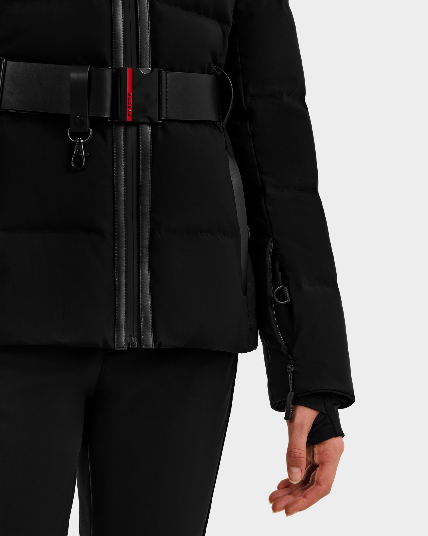 A close-up of a person wearing the Rudsak JOANNA - 8124788 Black women's down ski jacket with a wide removable belt, zipper details, and thumb hole in the sleeve, paired with black pants. The right hand's thumb is through the thumb hole.