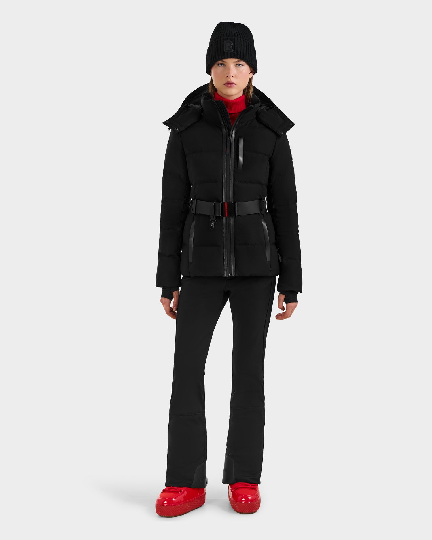 A person faces forward in the Rudsak JOANNA - 8124788 Black beanie and belted puffer jacket, paired with black pants and red shoes, set against a plain white background. Available for pre-order, shipping by November 30th.