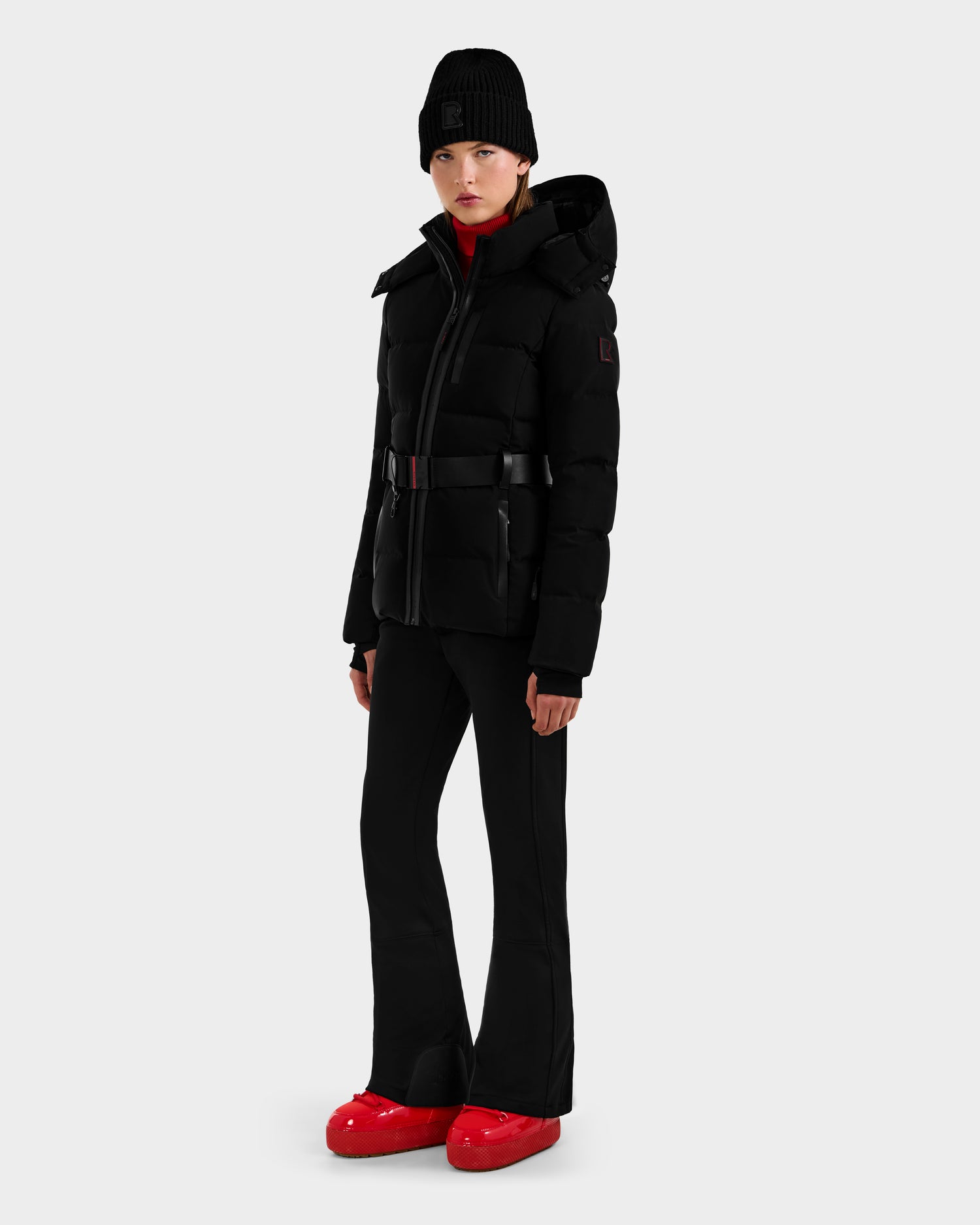 A person stands against a plain background wearing Rudsak's JOANNA - 8124788 Black women’s down ski jacket with removable belt, pairing it with black flared pants, a black beanie, and bright red shoes for a modern, winter-ready look.