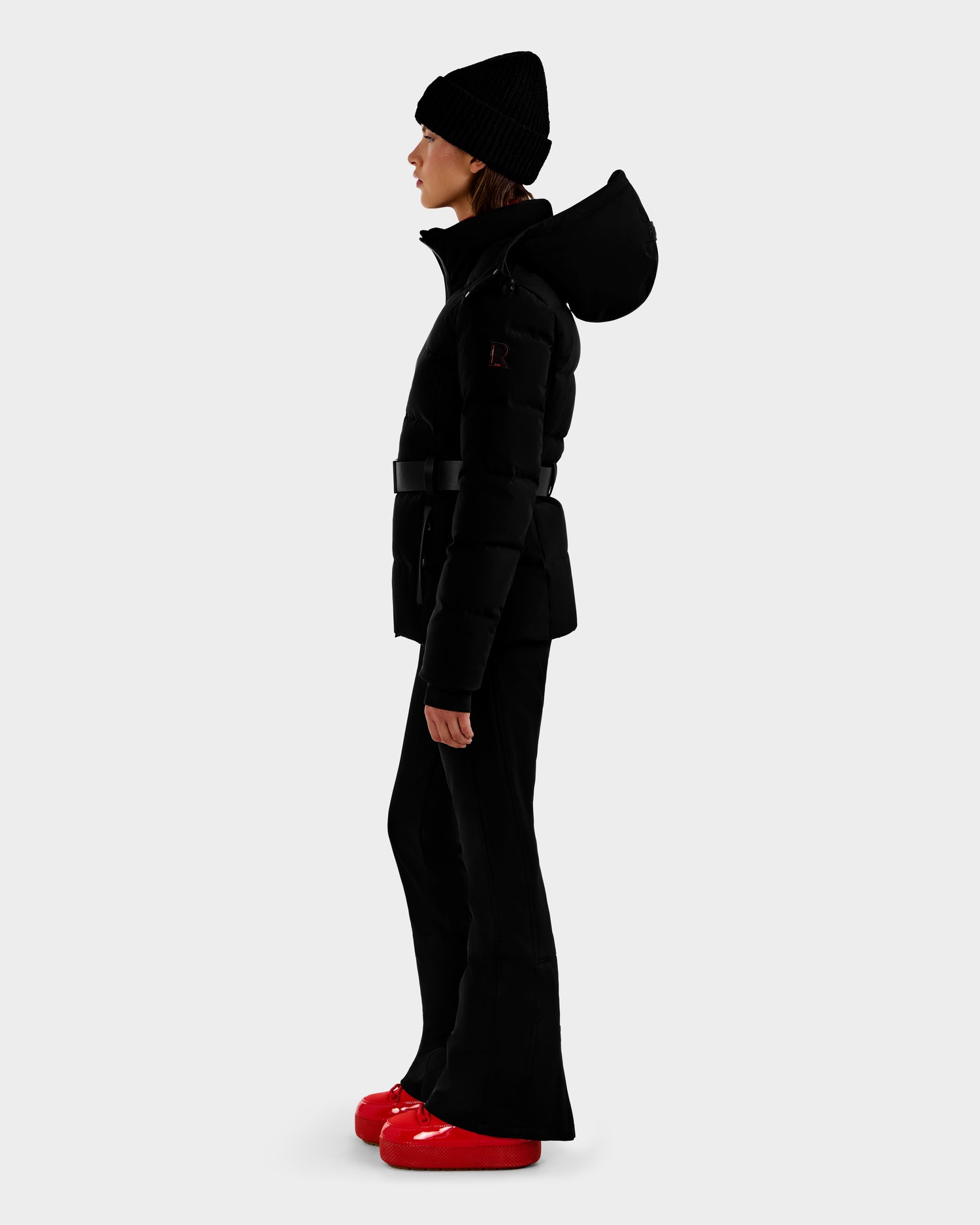 A person in profile wears the Rudsak JOANNA - 8124788 Black women’s down ski jacket with a hood, paired with a black beanie, black flared pants, and bright red puffy slippers against a plain light gray background.