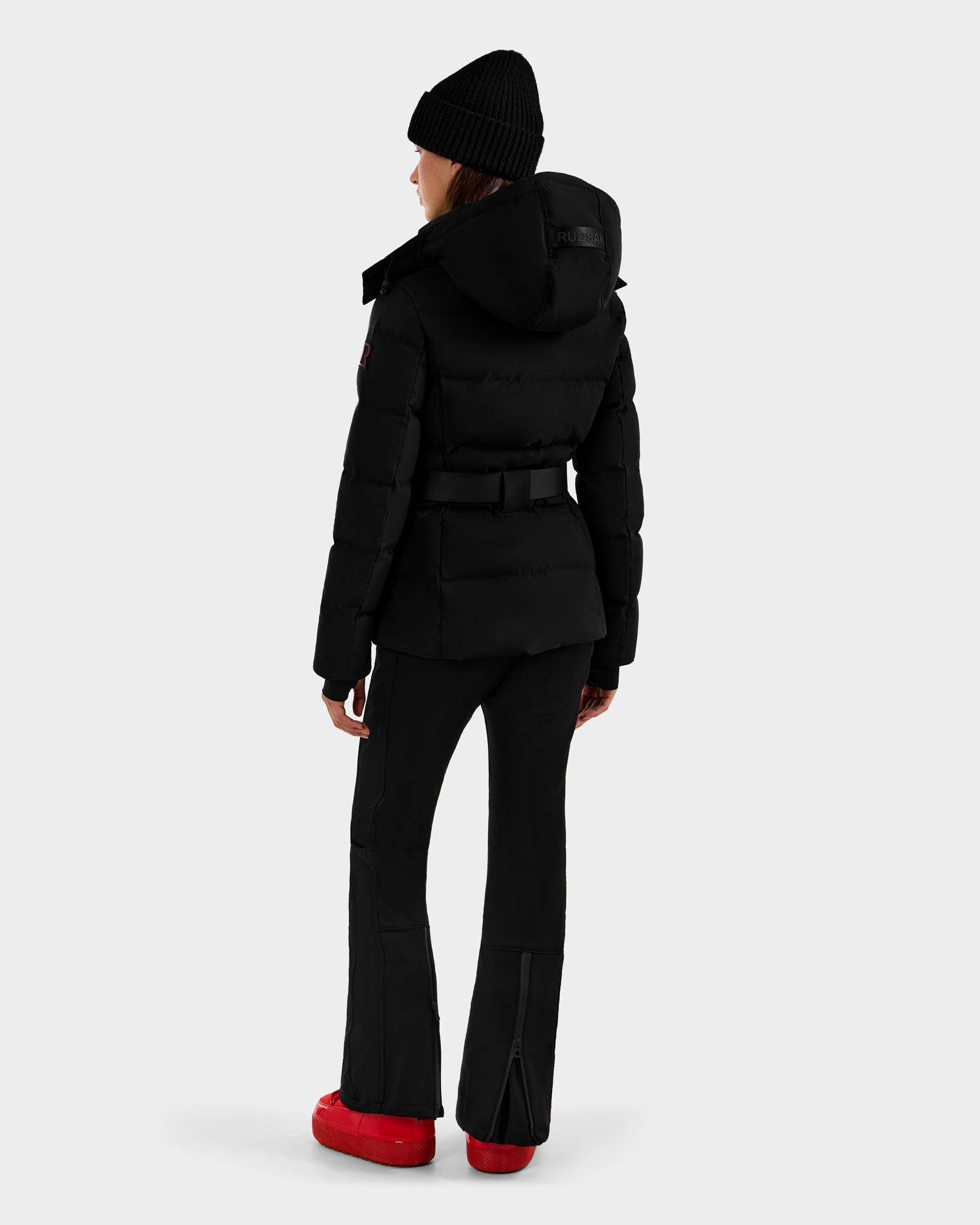 A person wearing the Rudsak JOANNA - 8124788 Black women's down ski jacket with a hood, black beanie, flared pants, and red boots stands facing away against a plain light gray background.