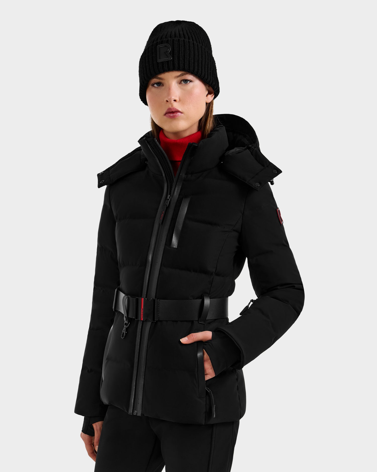 A woman wearing the Rudsak JOANNA - 8124788 Black women’s down ski jacket with a removable belt and black beanie stands against a light gray background, looking forward with a neutral expression.