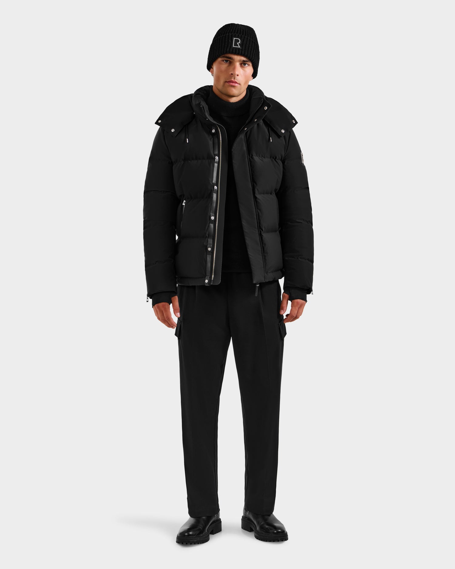 A man wears the Rudsak JOEL H - 6125582 Black men’s down parka, paired with a black beanie, turtleneck, pants, and shoes, standing against a plain white background—a true winter essential.