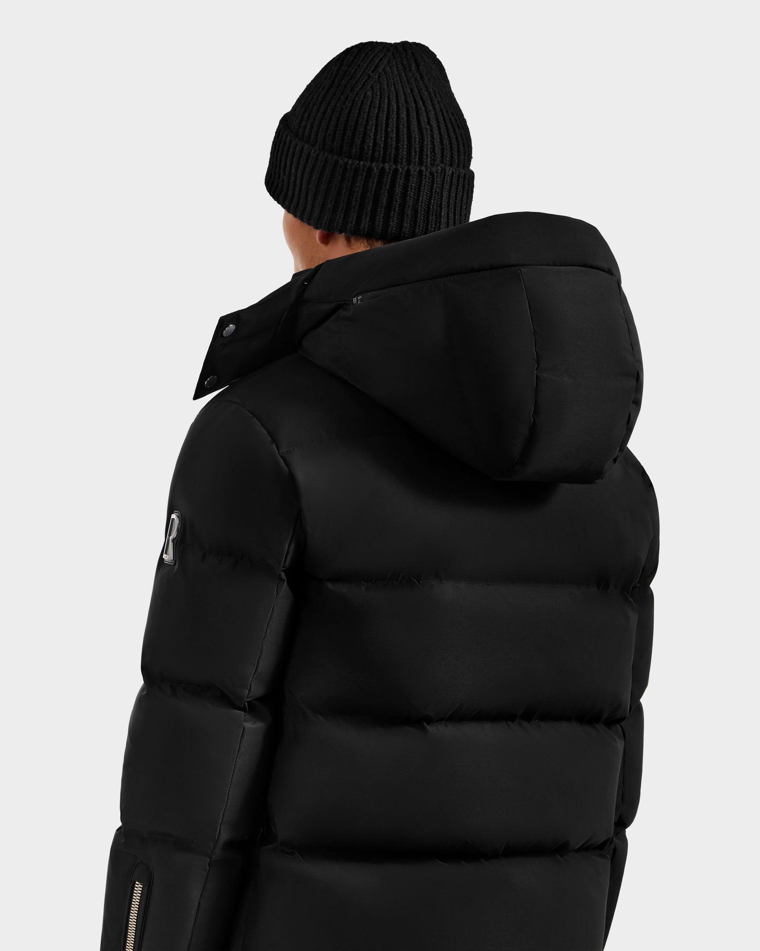 Wearing the Rudsak JOEL H - 6125582 Black RDS-certified down puffer jacket with hood and a black knitted beanie, this person is shown from behind against a light background—a true winter essential.