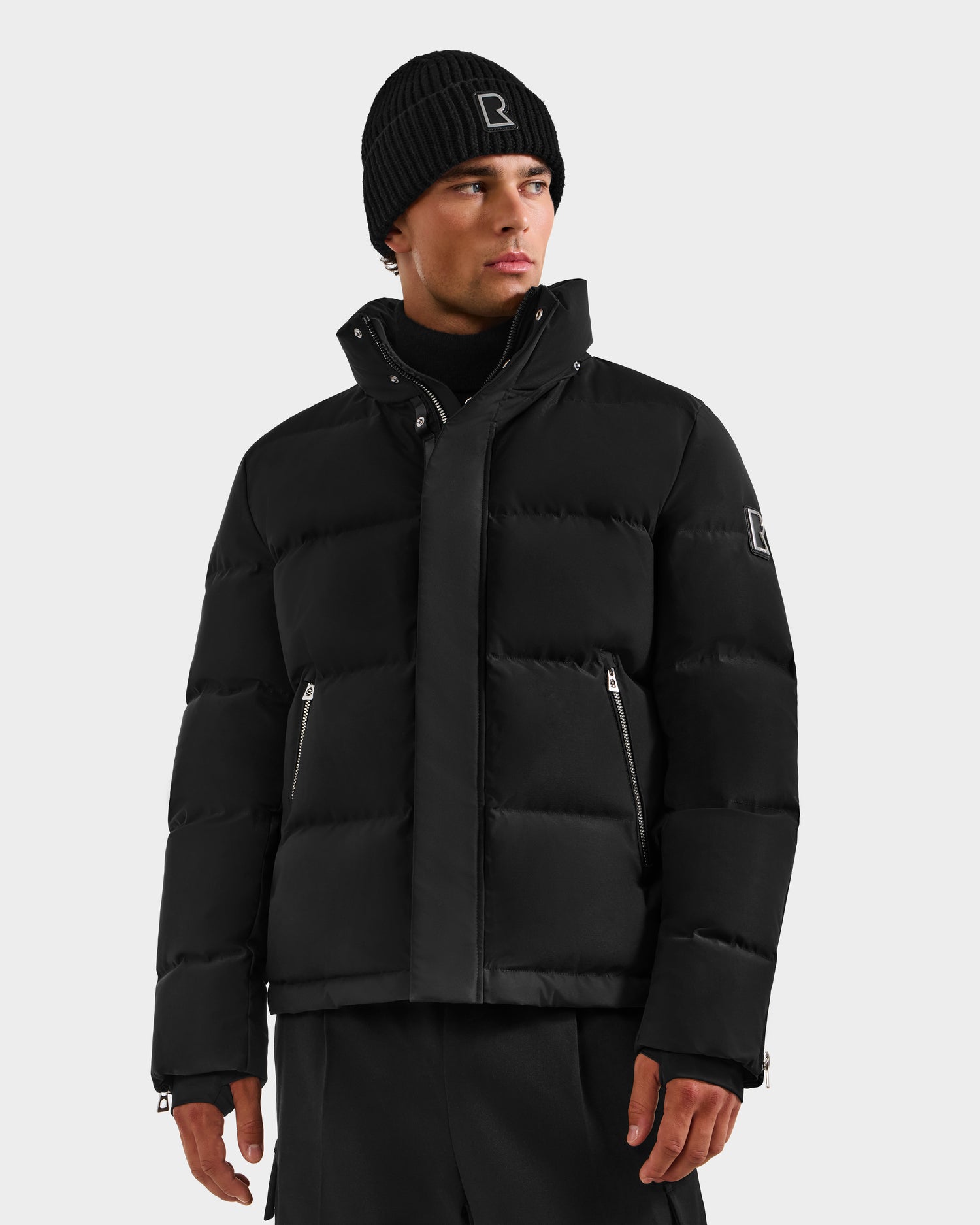 A man wears the Rudsak JOEL H - 6125582 Black down parka with a black beanie and pants, standing against a light gray background with a neutral expression.