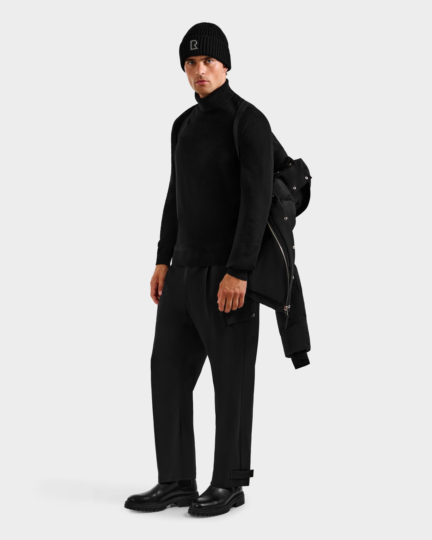 A man dressed in black stands against a plain light background, holding the Rudsak JOEL H - 6125582 Black men’s down parka—an essential winter piece—over his shoulder.