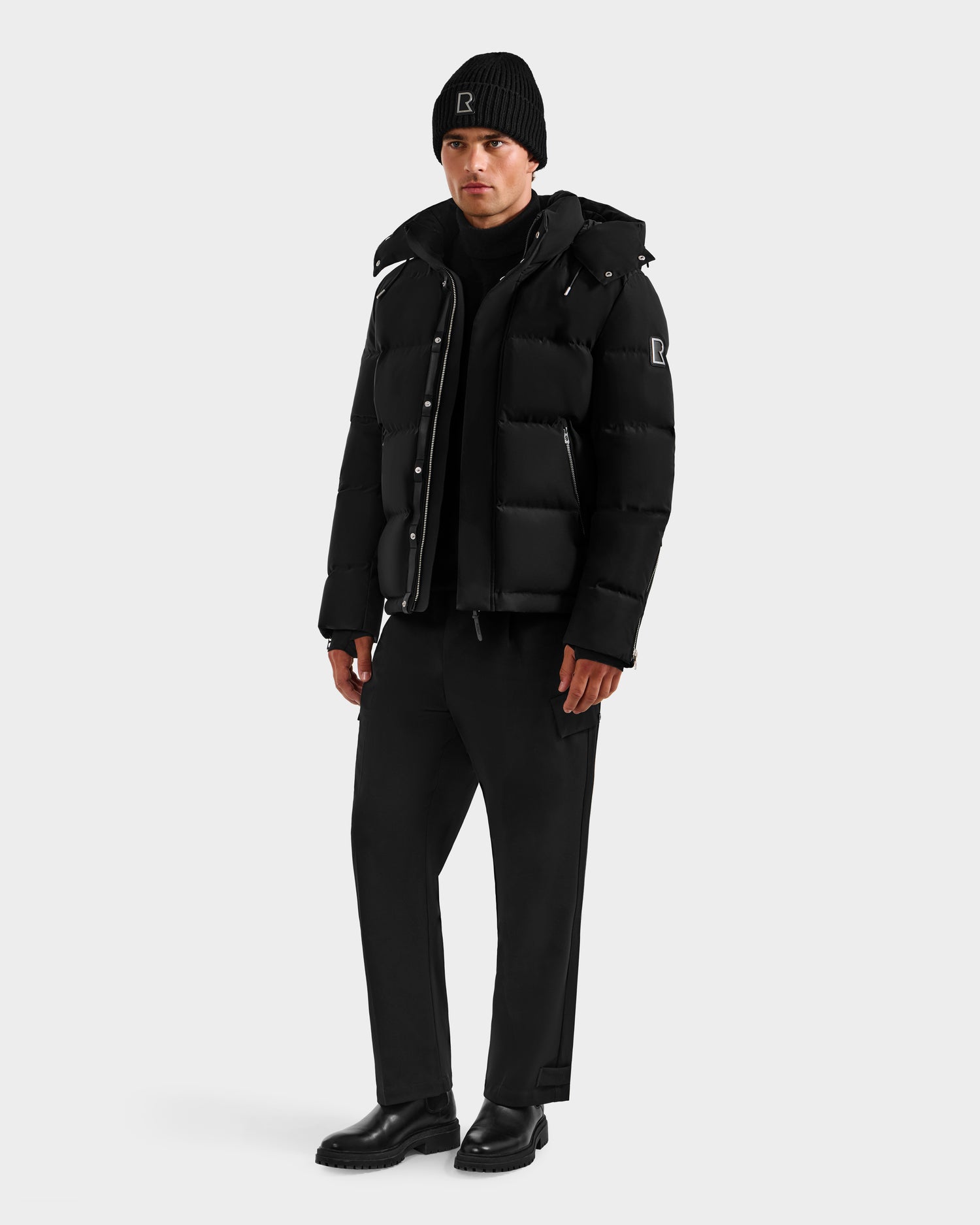 A person wears Rudsak’s JOEL H - 6125582 Black men’s down parka with RDS-certified down, styled with black beanie, shirt, pants, and boots, set against a plain light-gray background for a sleek winter look.