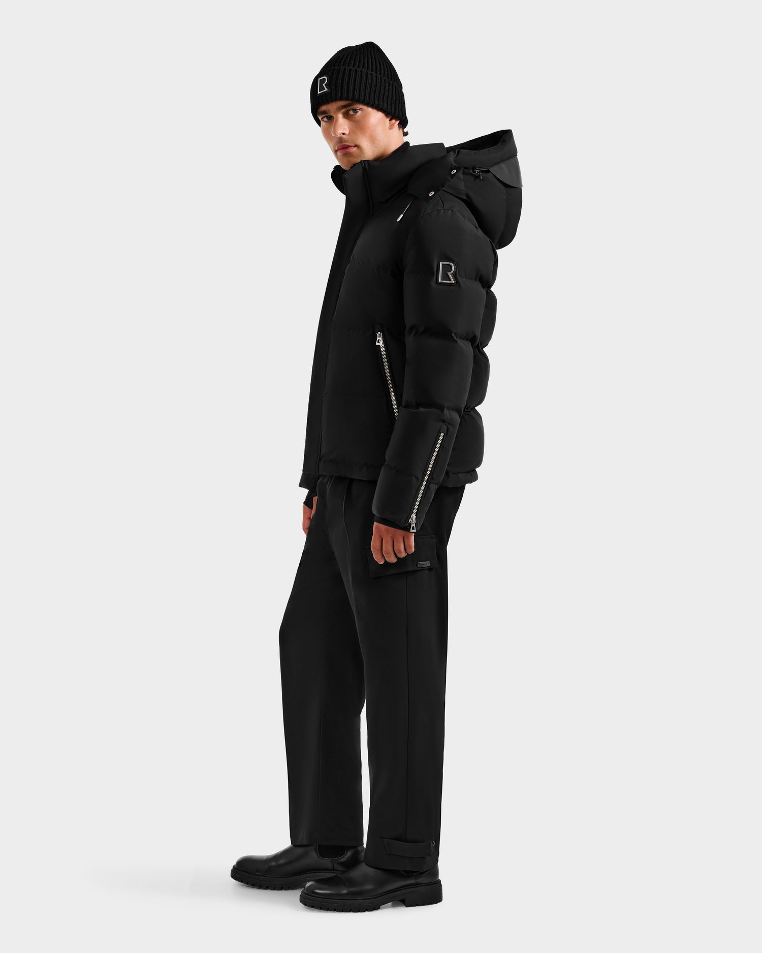 A person wearing the Rudsak JOEL H - 6125582 Black men’s down parka with RDS-certified down, paired with a black beanie, pants, and boots, stands slightly to the side against a plain light background—a true winter essential.