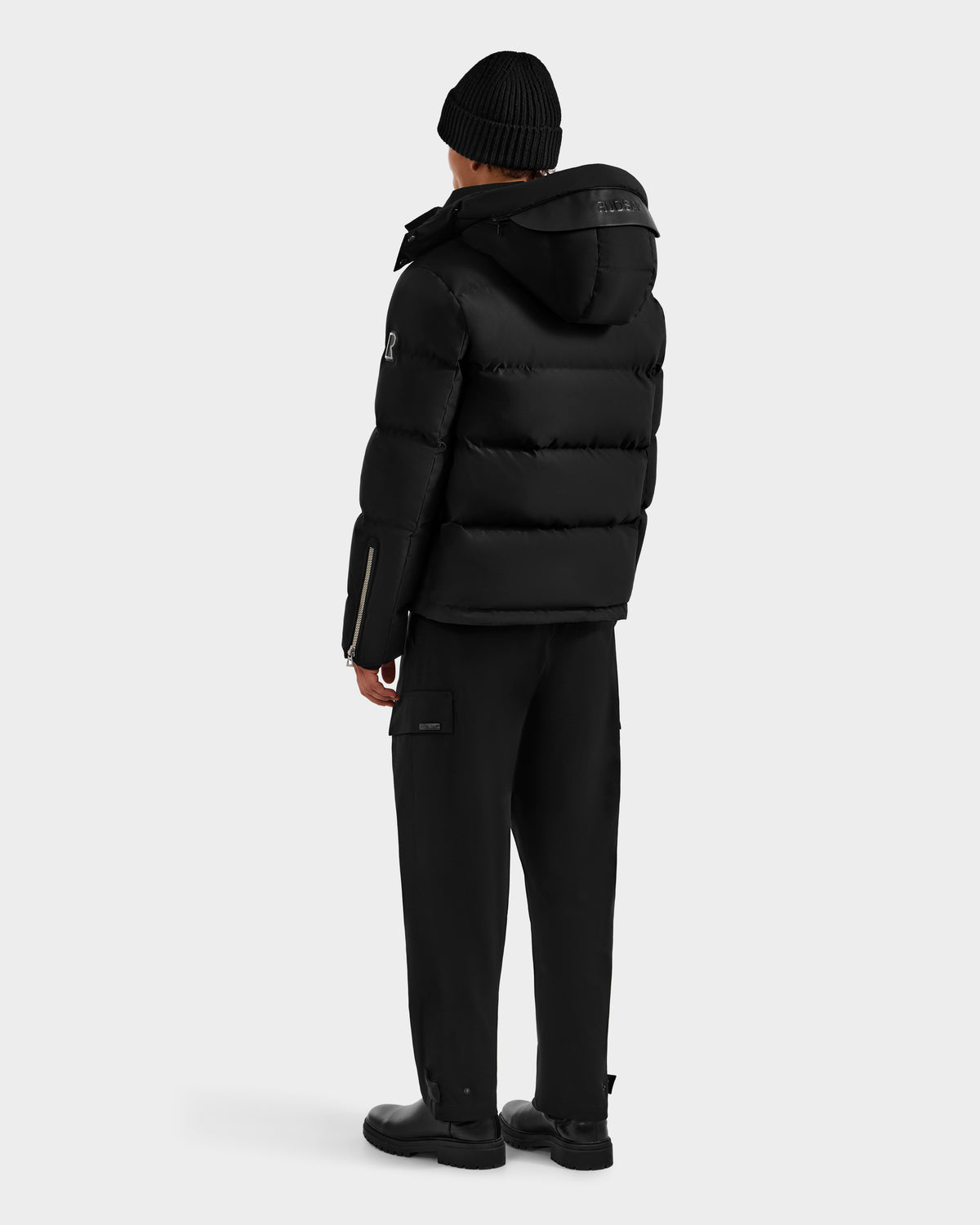 A person faces away wearing Rudsak’s JOEL H - 6125582 Black men’s down parka with a hood, paired with black pants, boots, and a black knit beanie—a true winter essential—set against a plain light background.