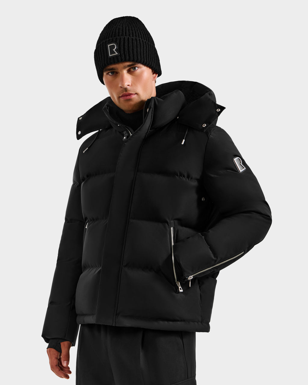 A man wears the Rudsak JOEL H - 6125582 Black men’s down parka with a hood and silver zippers, paired with a black beanie featuring a white letter R, standing against a plain light background—a true winter essential.