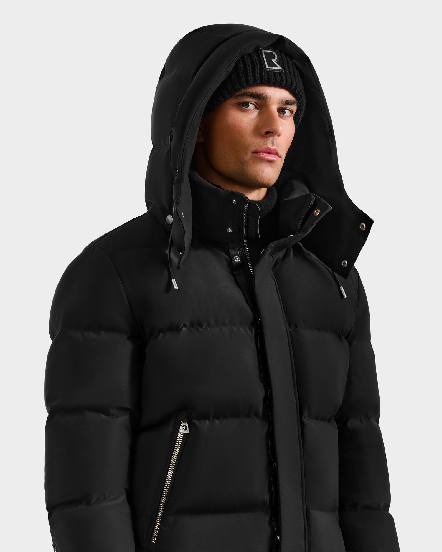 A man wears the Rudsak JOEL H - 6125582 Black men’s down parka with the hood up and a black knit beanie, posing against a plain light background—an ideal winter essential.