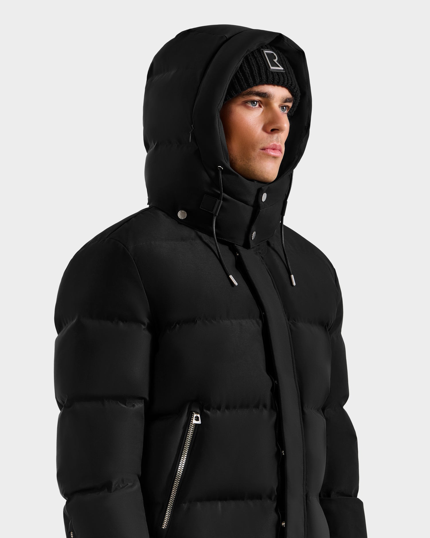 A man wears the Rudsak JOEL H - 6125582 Black men’s down parka with hood and zipper details, paired with a black beanie featuring a small white logo, against a plain light background—showcasing an RDS-certified winter essential.
