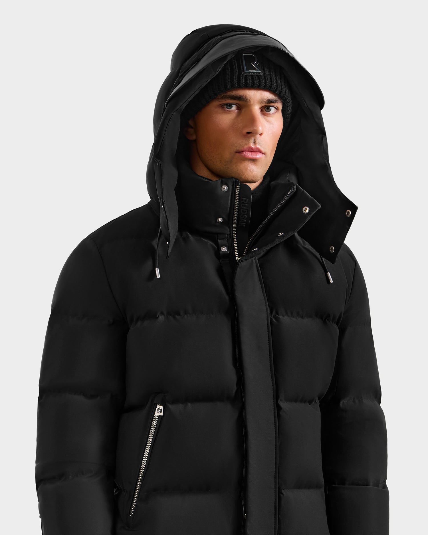 A man wearing the Rudsak JOEL H - 6125582 Black RDS-certified down puffer jacket with a hood and a black beanie, standing against a plain light grey background—a true winter essential.