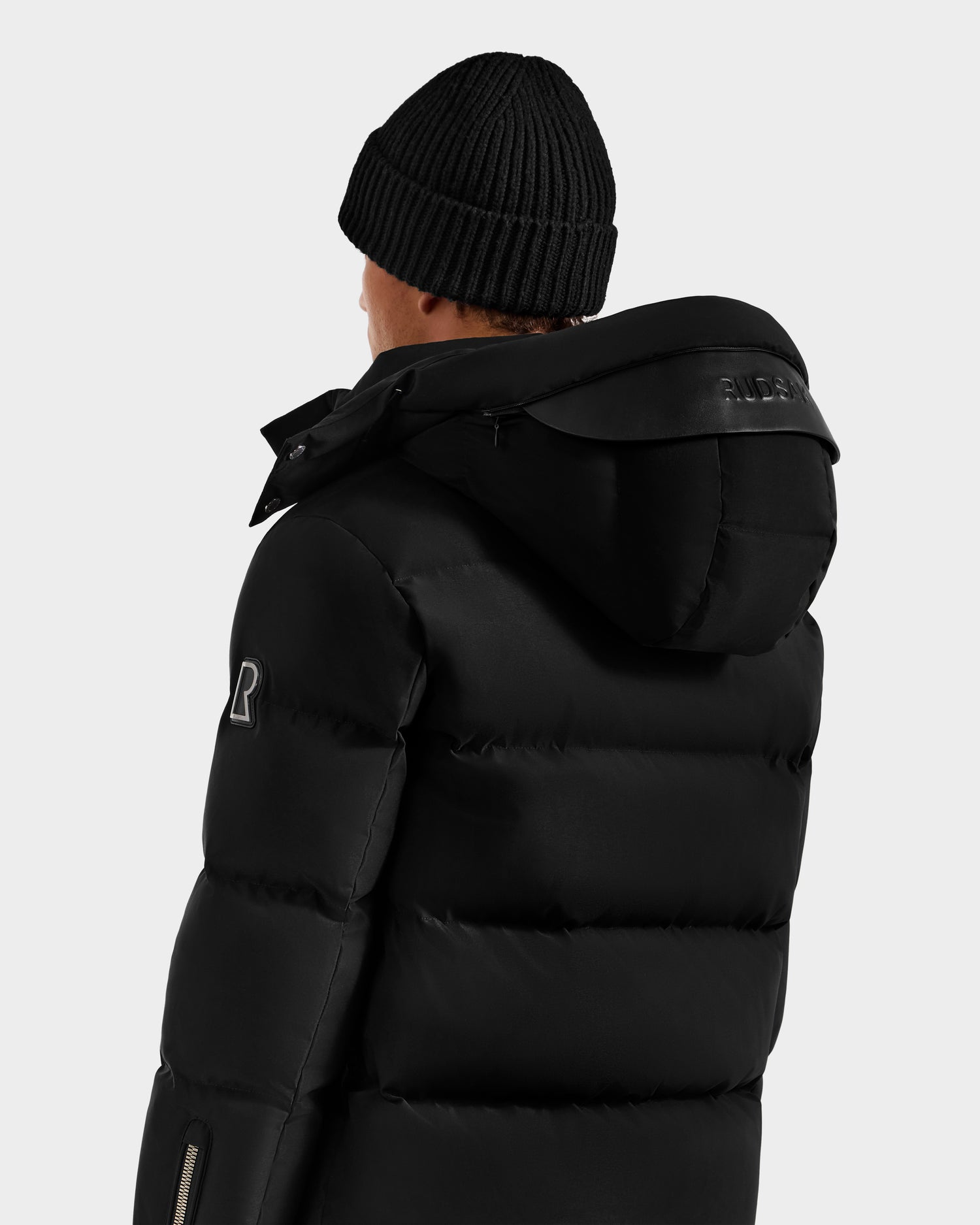 A person wearing the Rudsak JOEL H - 6125582 Black RDS-certified down puffer jacket with hood and a black knit beanie, viewed from the back against a plain light background—a true winter essential.