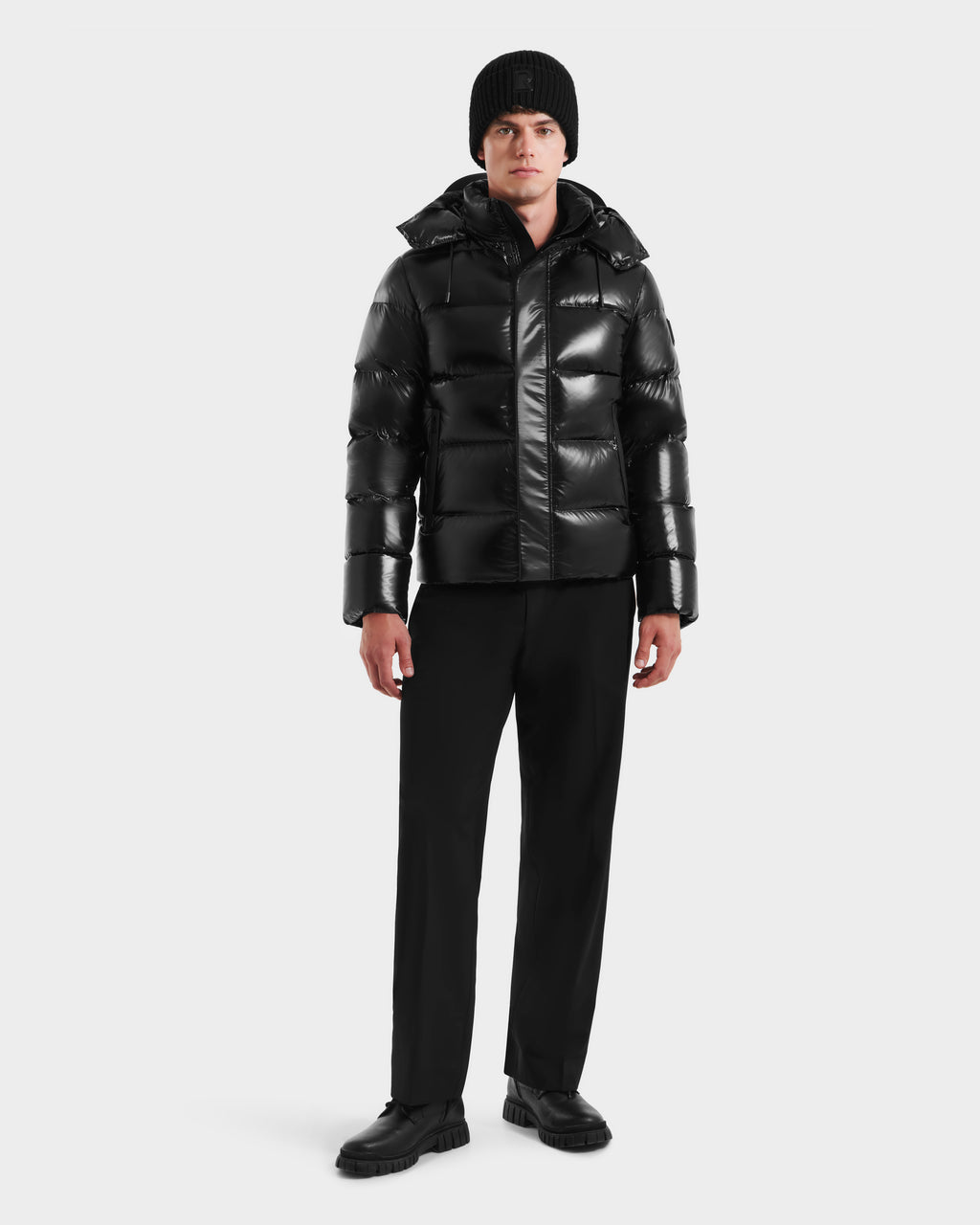 A man stands facing forward against a white background, wearing the Rudsak JOEL S - 6124606 Black down puffer jacket with black pants, shoes, and a knit beanie, highlighting stylish winter outerwear.
