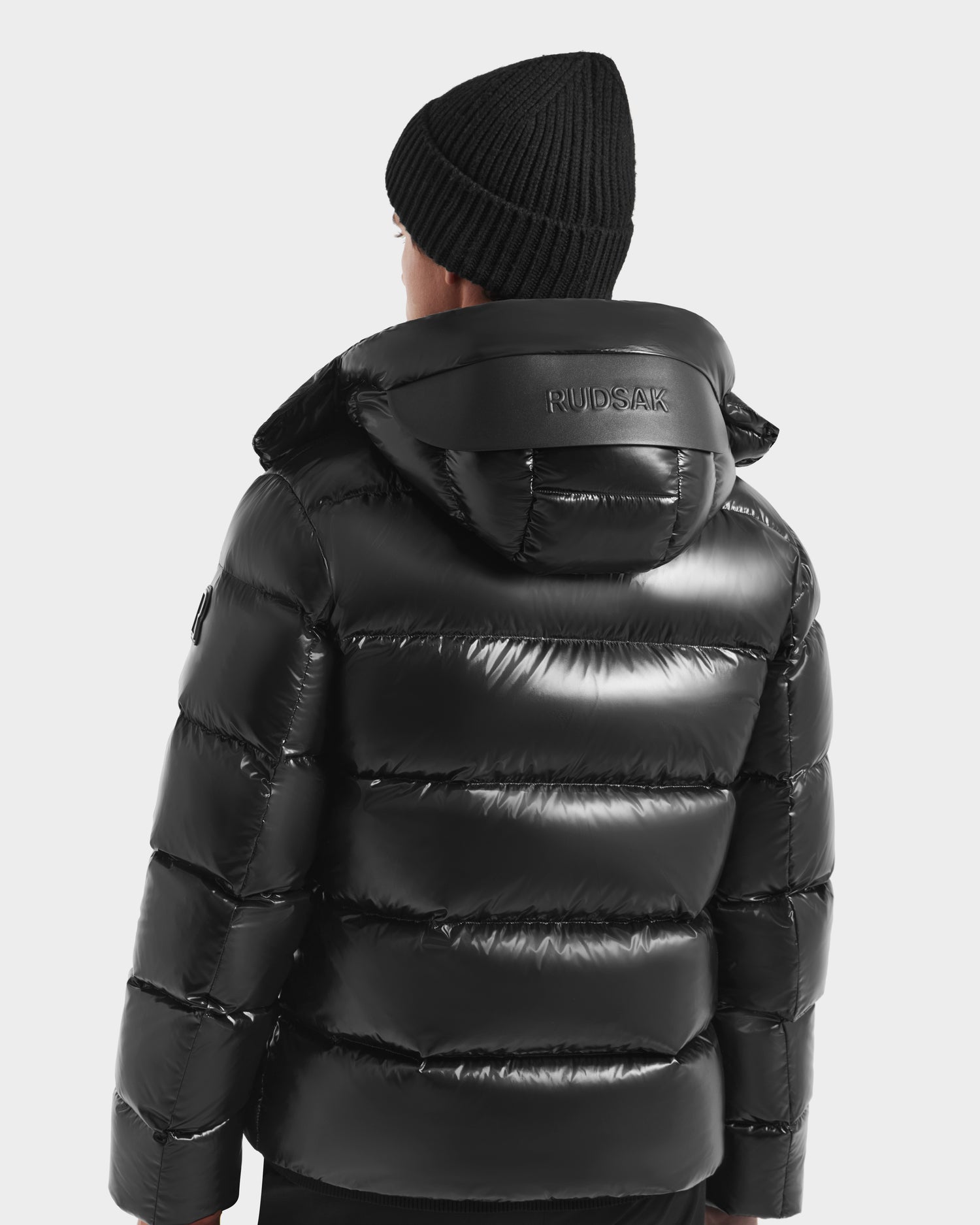 A person in the Rudsak JOEL S - 6124606 Black shiny down puffer jacket with hood and a black knit beanie is shown from the back, “RUDSAK” visible on the collar, showcasing premium winter outerwear style.