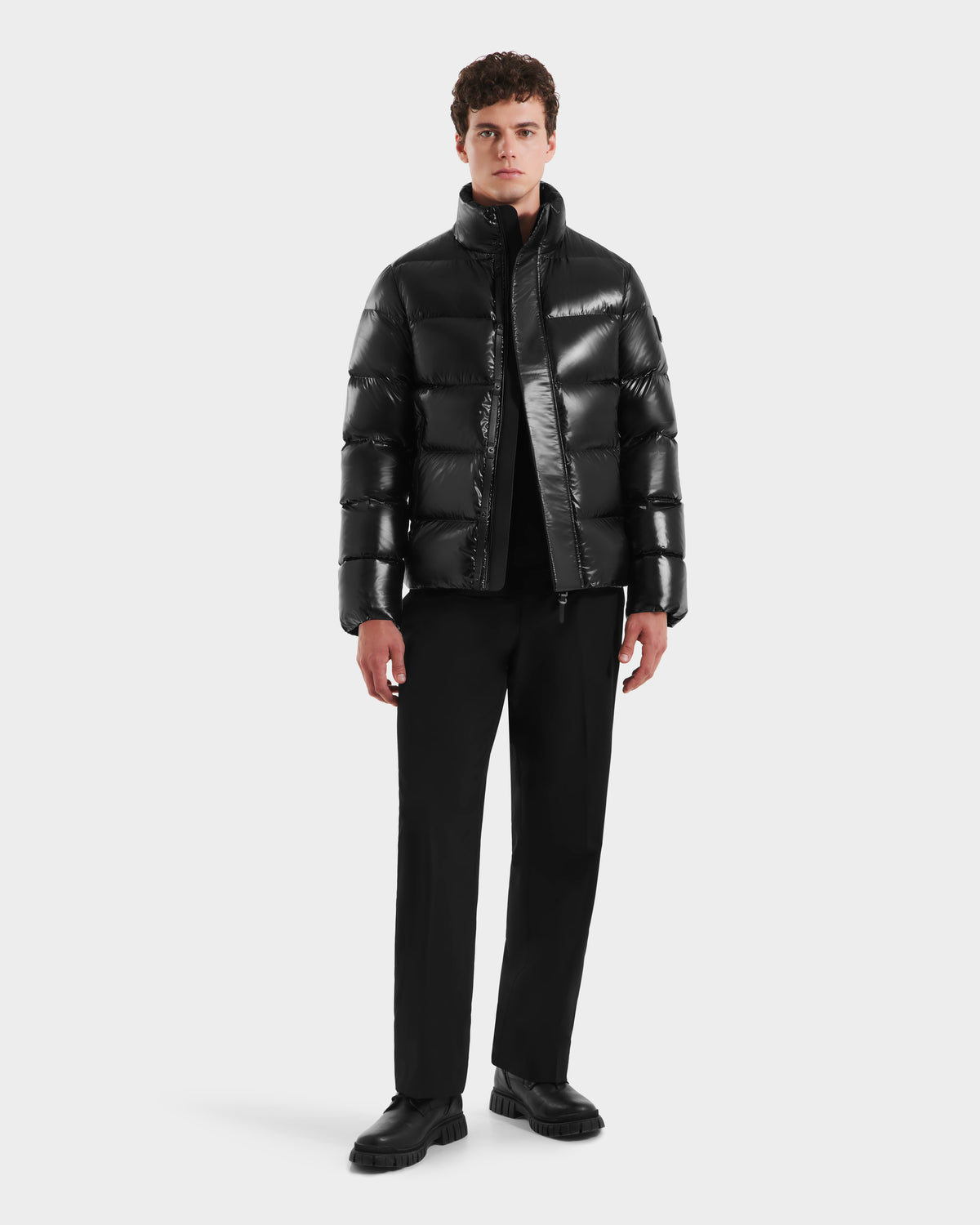 A man stands facing forward in a Rudsak JOEL S - 6124606 Black shiny down puffer jacket over a black shirt, paired with black pants and shoes. A light background highlights his stylish winter outerwear.