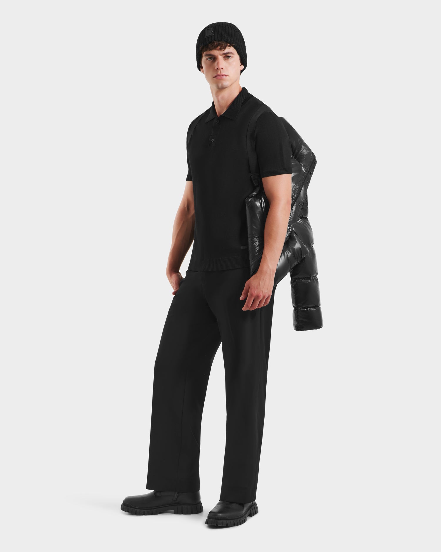 A man stands against a plain background wearing a black knit beanie, polo, pants, and shoes, with the Rudsak JOEL S - 6124606 Black premium down puffer jacket draped over his shoulder for an effortless winter look.
