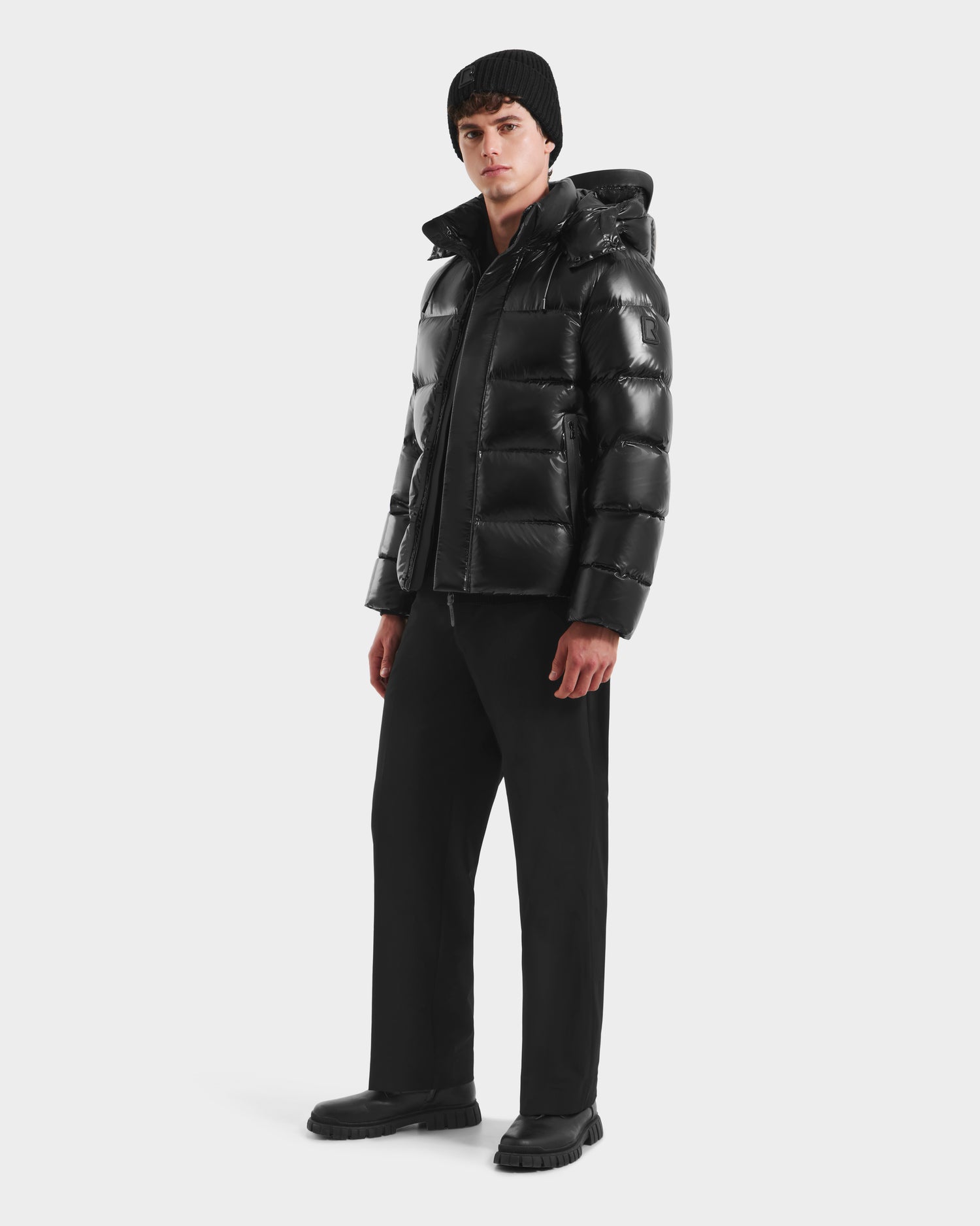 A person wearing the Rudsak JOEL S - 6124606 Black down puffer jacket, paired with black pants, boots, and a beanie, stands against a plain white background.