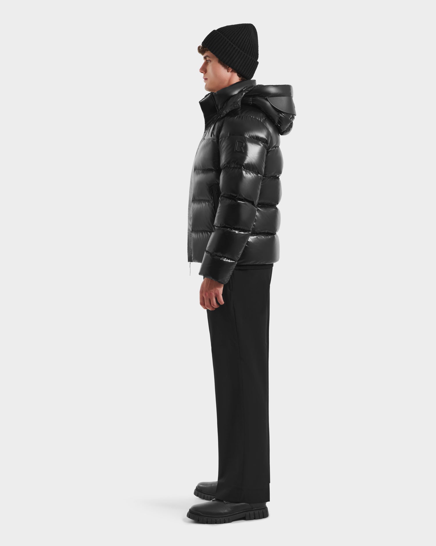 A person stands in profile against a plain white background wearing the Rudsak JOEL S - 6124606 Black premium down puffer jacket, paired with a black beanie, black pants, and black shoes.