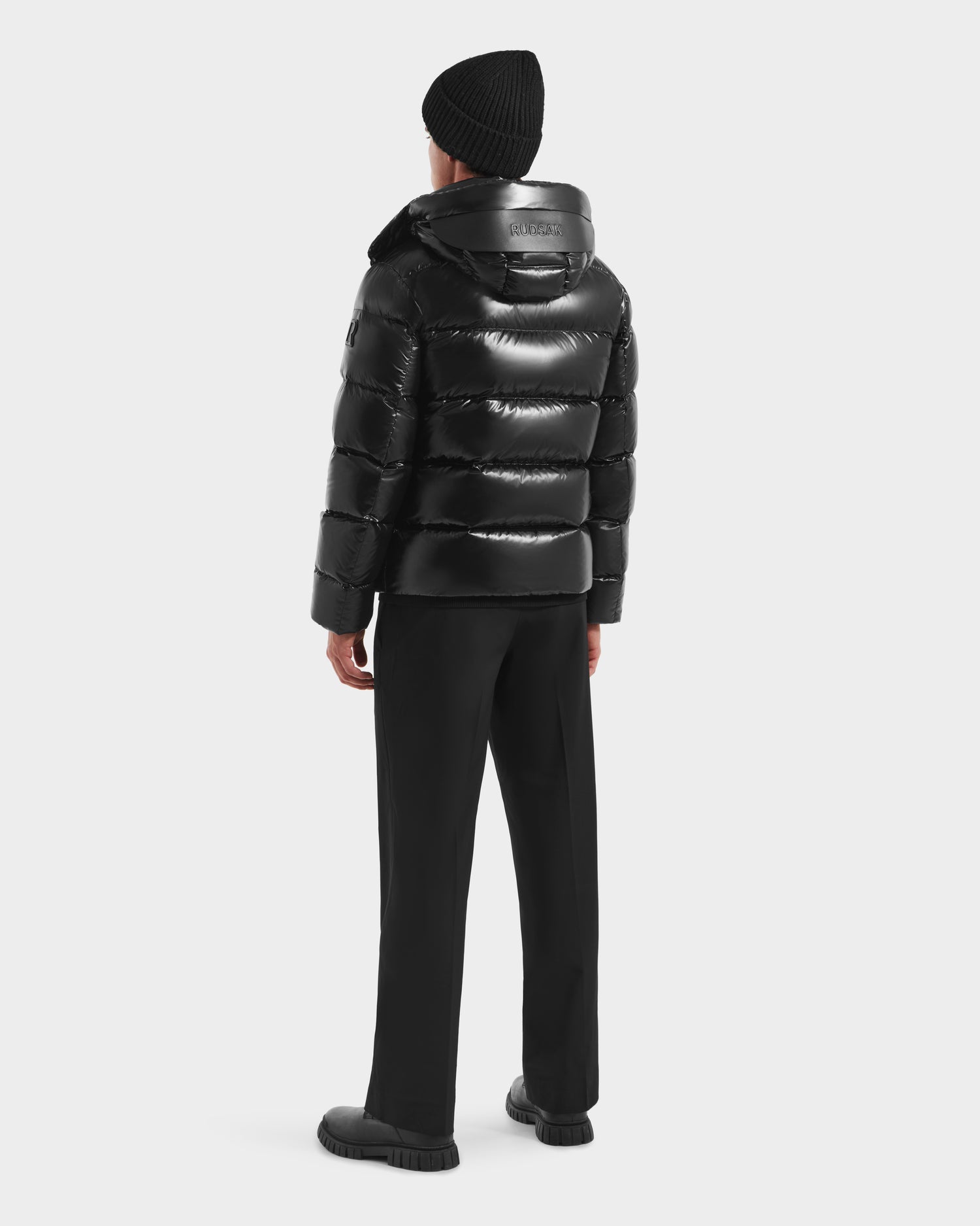 A person faces away, wearing the Rudsak JOEL S - 6124606 Black down puffer jacket with a hood, paired with black pants and shoes and a black knit beanie against a light background—showcasing sleek winter style.