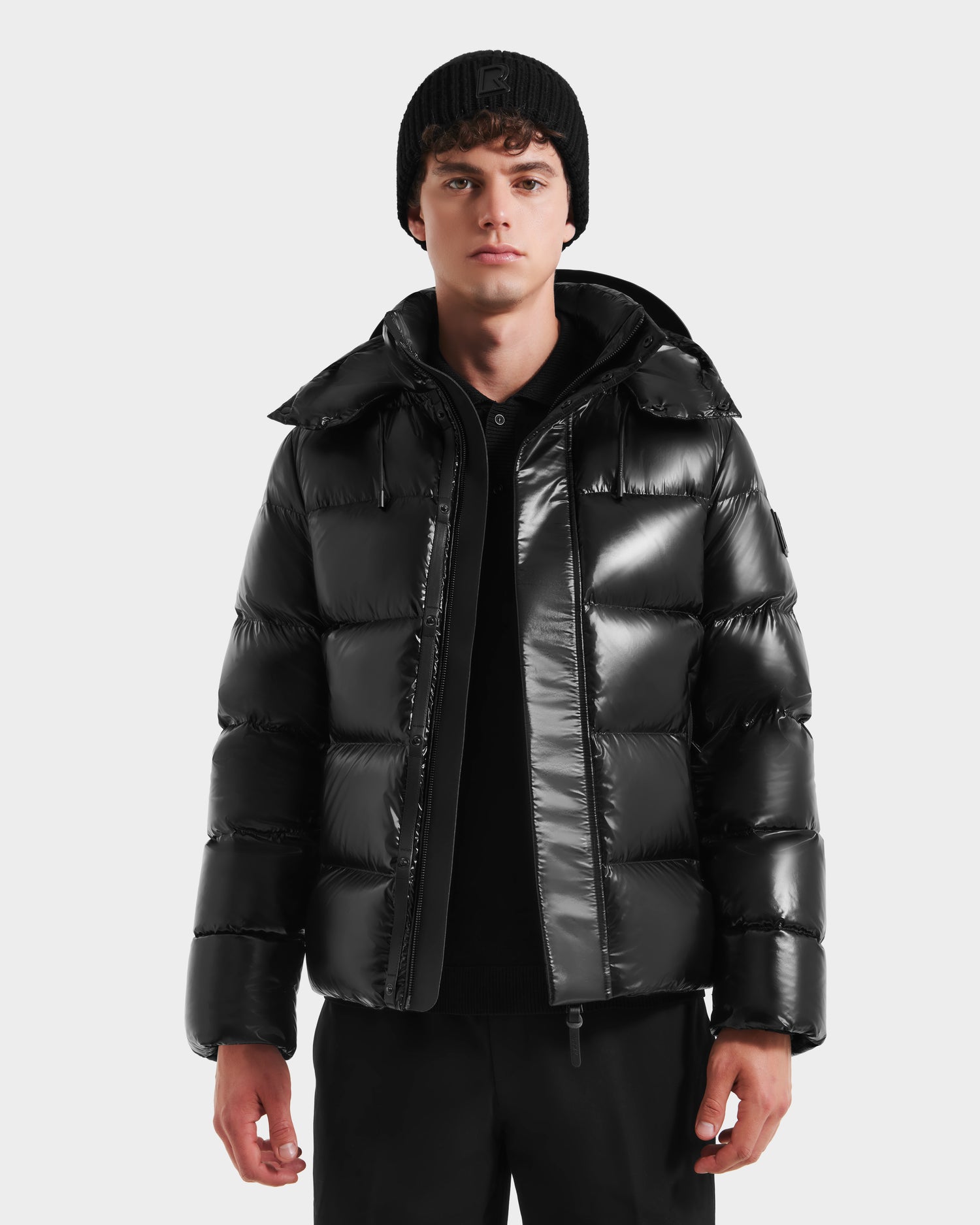 A young man wears the Rudsak JOEL S - 6124606 Black premium down puffer jacket, paired with a black beanie and pants, standing facing forward against a plain light gray background.