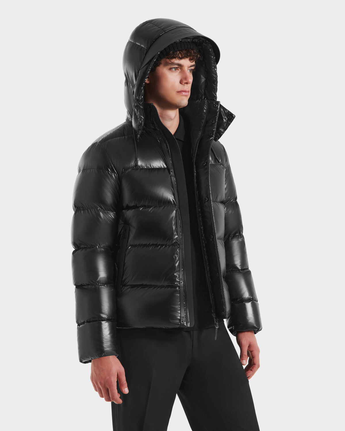 A young man with curly hair wears the Rudsak JOEL S - 6124606 Black premium down puffer jacket with a hood, paired with dark pants and a black shirt, standing against a plain white background.