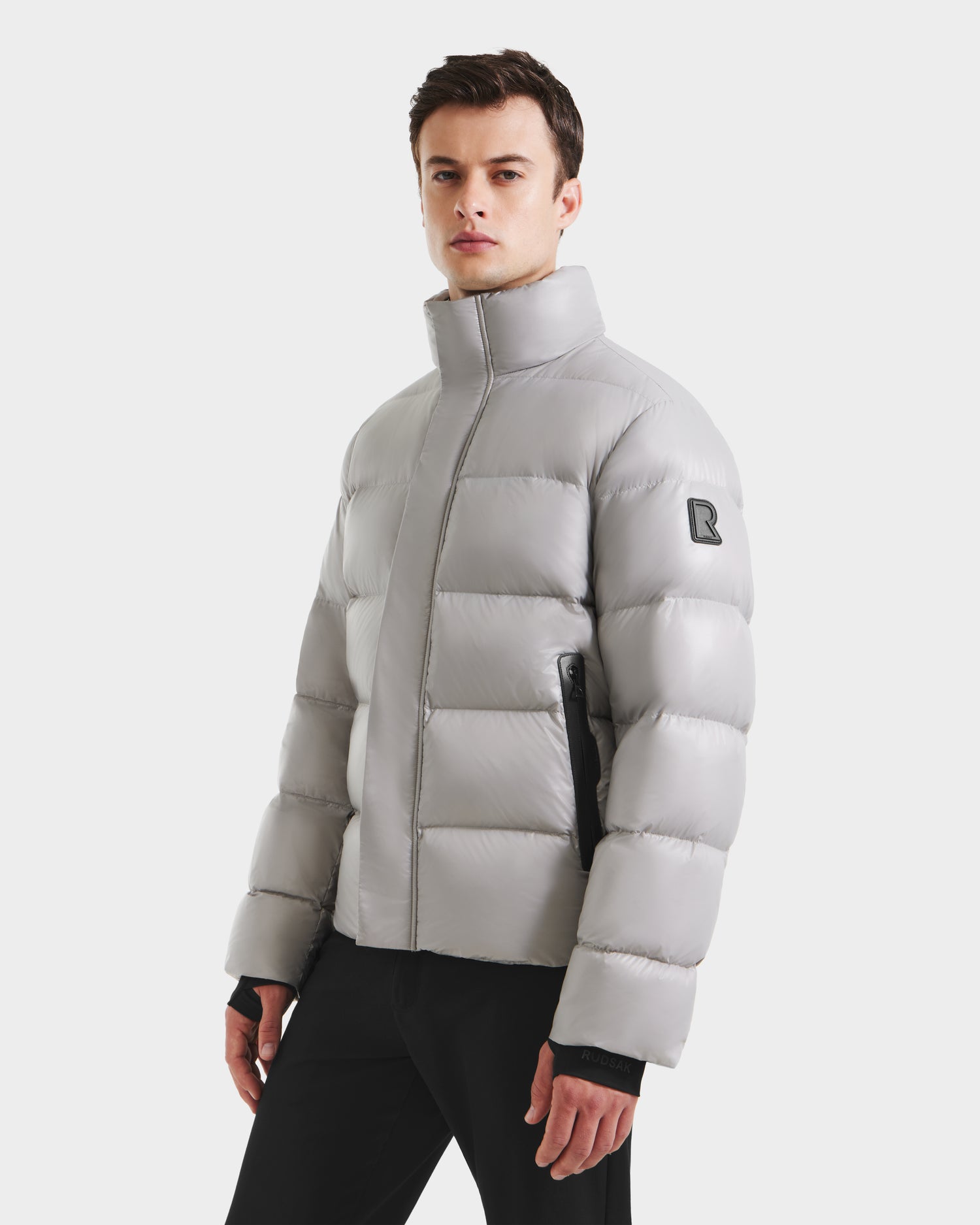 A man stands against a white background wearing the Rudsak JOEL S - 6124606 Light Grey down puffer jacket with a high collar, black zippers, and a sleeve patch, paired with black pants—perfect for winter.