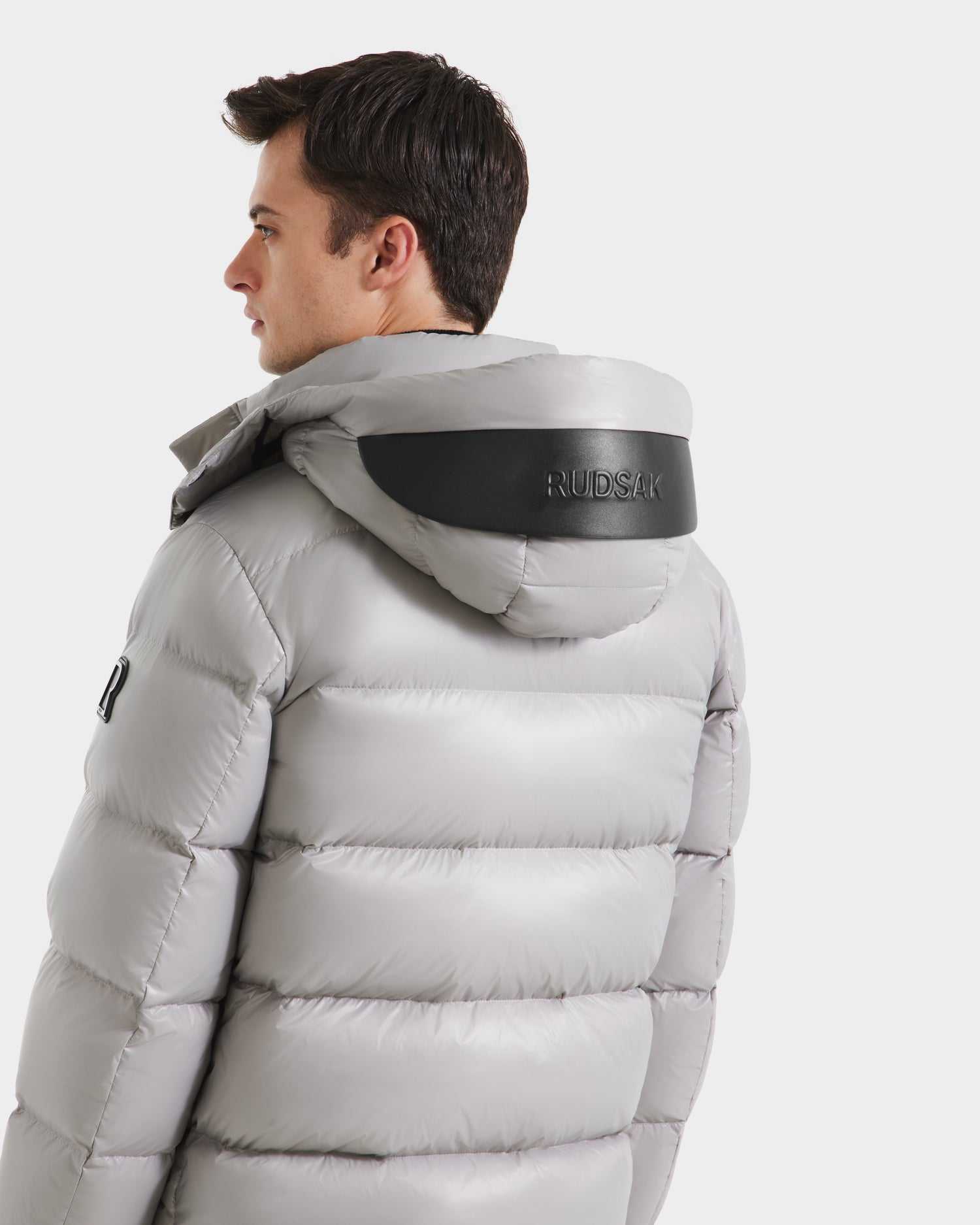 A man faces away, looking left, in the Rudsak JOEL S - 6124606 light grey down puffer jacket with a hood featuring a black panel embossed with the brand name.