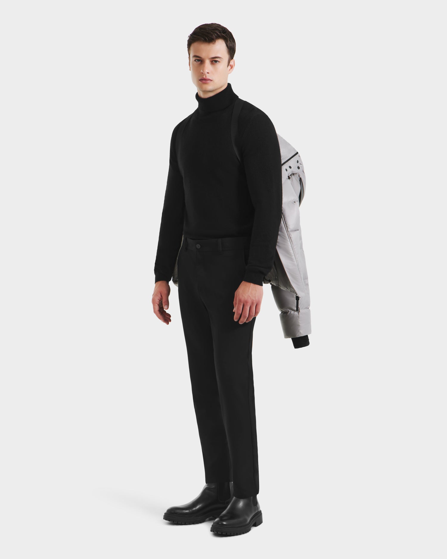 A man stands facing forward against a white background, wearing black clothes and boots, with the Rudsak JOEL S - 6124606 Light Grey premium down jacket draped over his left shoulder.