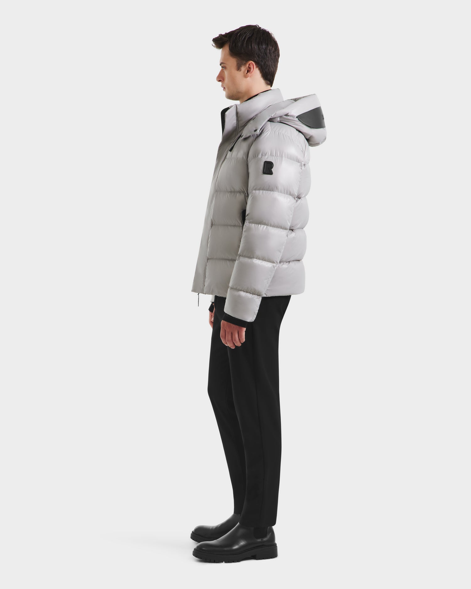 A man stands in profile wearing the Rudsak JOEL S - 6124606 Light Grey down puffer jacket with a hood, paired with black pants and boots, set against a plain white background.