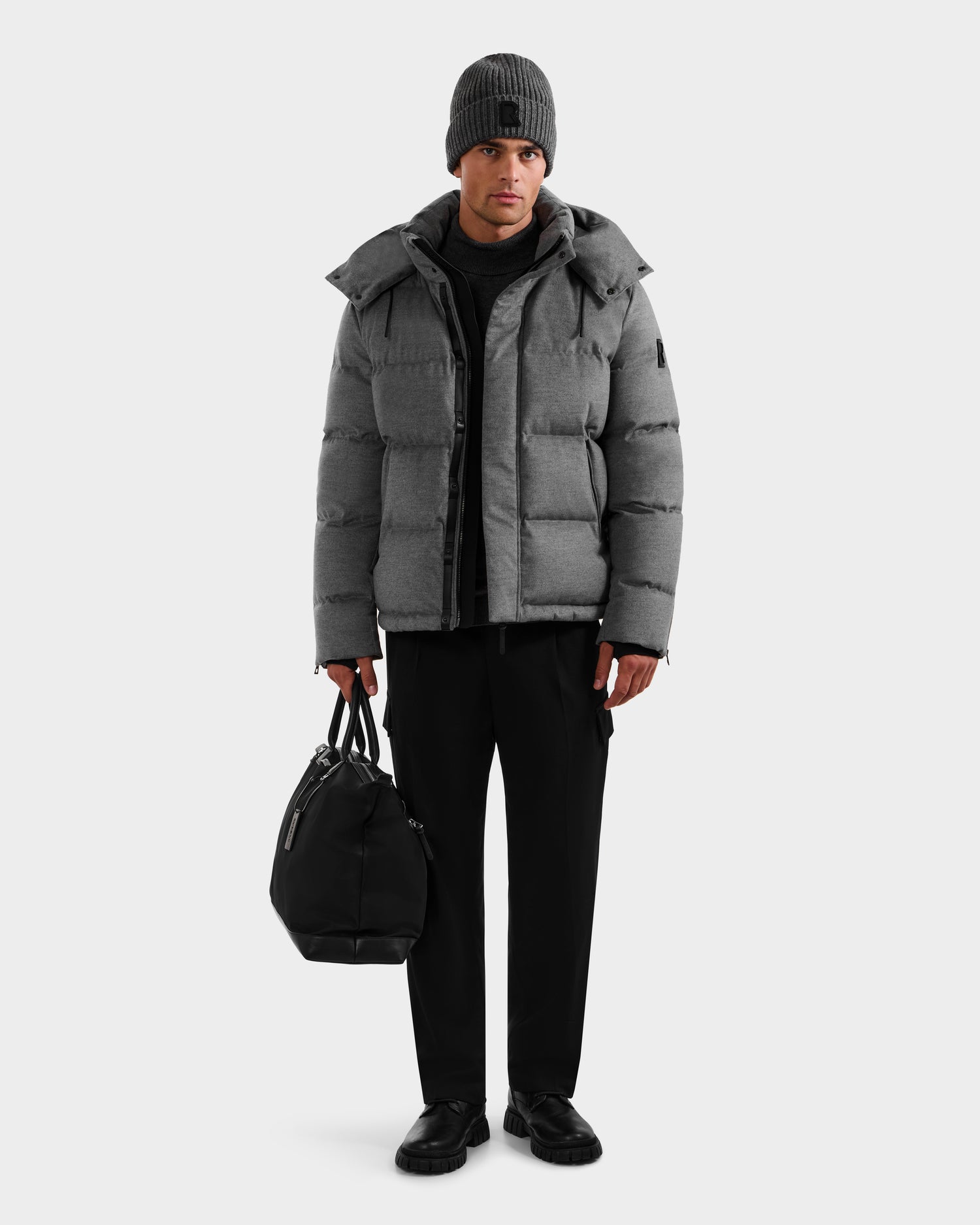 A man stands against a plain light background wearing the Rudsak JOEL W - 6125683 Heather Charcoal men’s puffer jacket with removable hood, paired with black pants, black shoes, a gray knit beanie, and holding a black duffel bag.