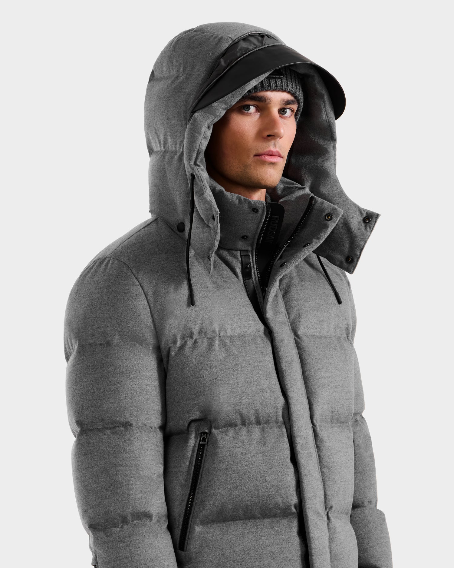 Wearing the Rudsak JOEL W - 6125683 Heather Charcoal men’s wool-blend down puffer with black zipper and hood, a gray knit beanie, and black visor cap, a person looks to the side against a light background.