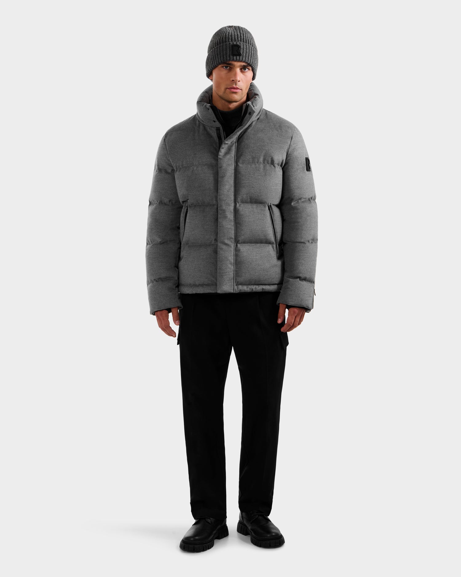 A man faces forward wearing the Rudsak JOEL W - 6125683 Heather Charcoal down jacket with removable hood, paired with black pants, black shoes, and a gray knit beanie against a plain light background.