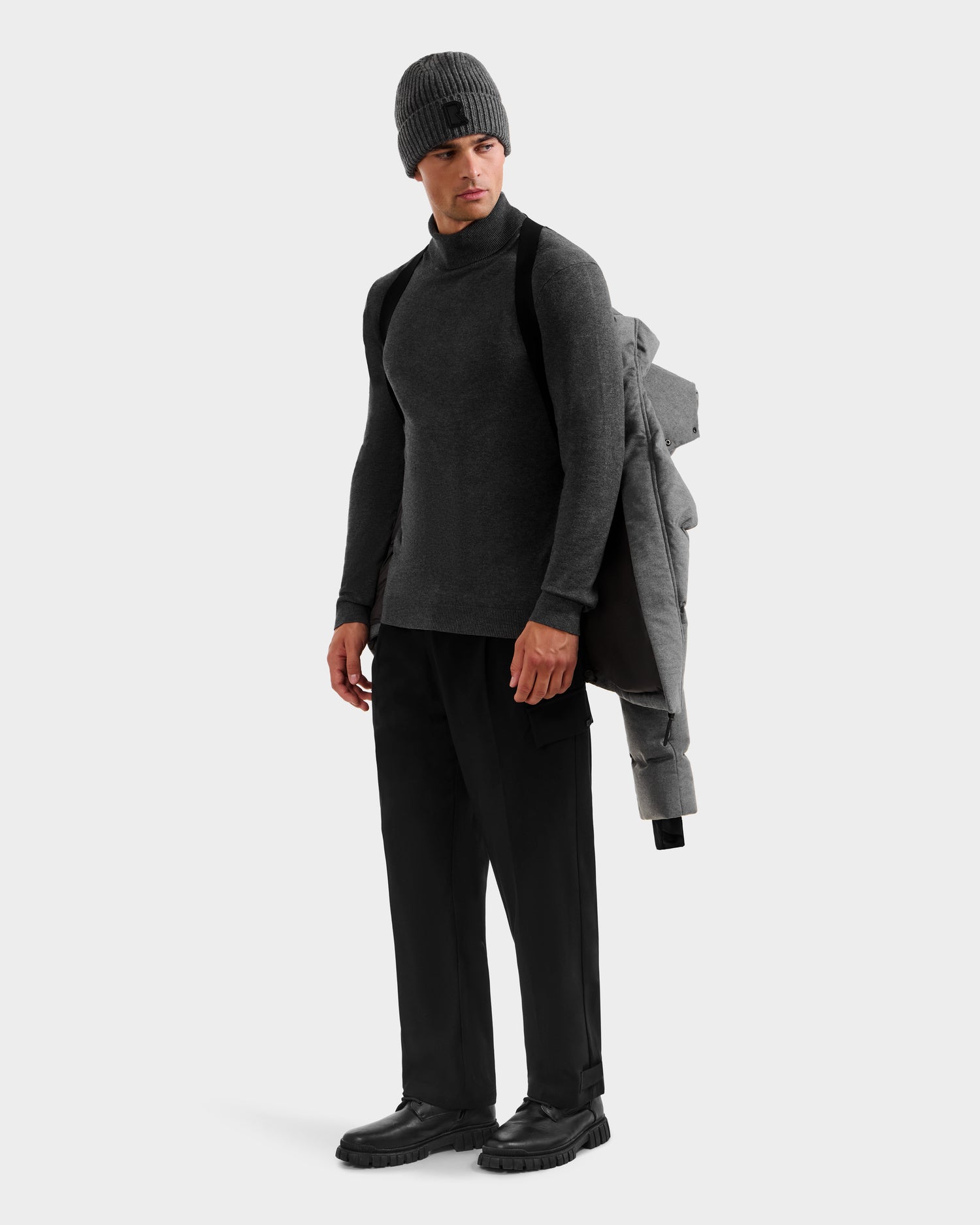 A man in a gray knit beanie, dark gray long-sleeve shirt, black pants, and shoes stands with the Rudsak JOEL W - 6125683 Heather Charcoal wool-blend down puffer draped over his shoulder and a black backpack, looking sideways against a plain background.