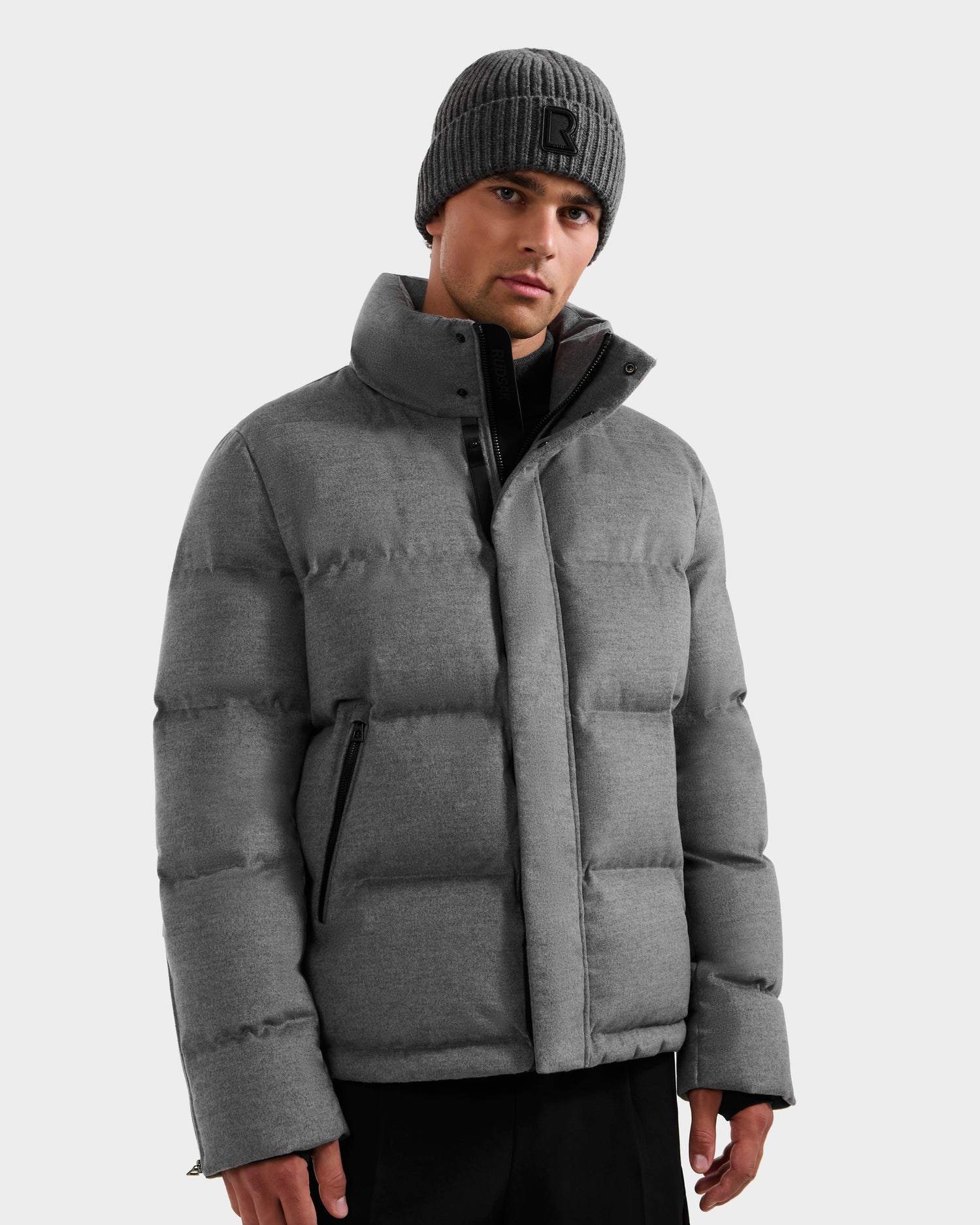 A man wears the Rudsak JOEL W - 6125683 Heather Charcoal wool-blend down puffer with removable hood, paired with black pants and a gray knit beanie, standing against a plain light gray background with a neutral expression.