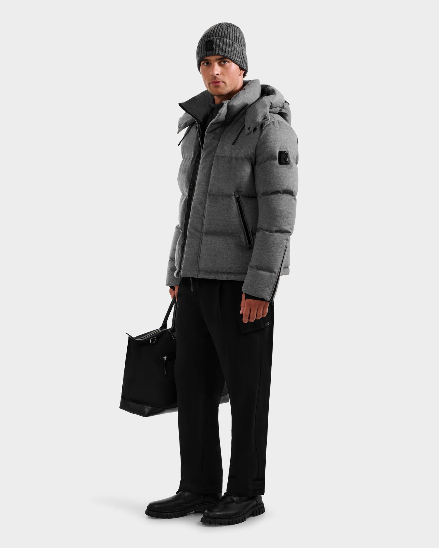 A man in the Rudsak JOEL W - 6125683 Heather Charcoal premium down jacket with a removable hood, black pants, shoes, and a gray knit beanie stands holding a black bag against a plain light background.