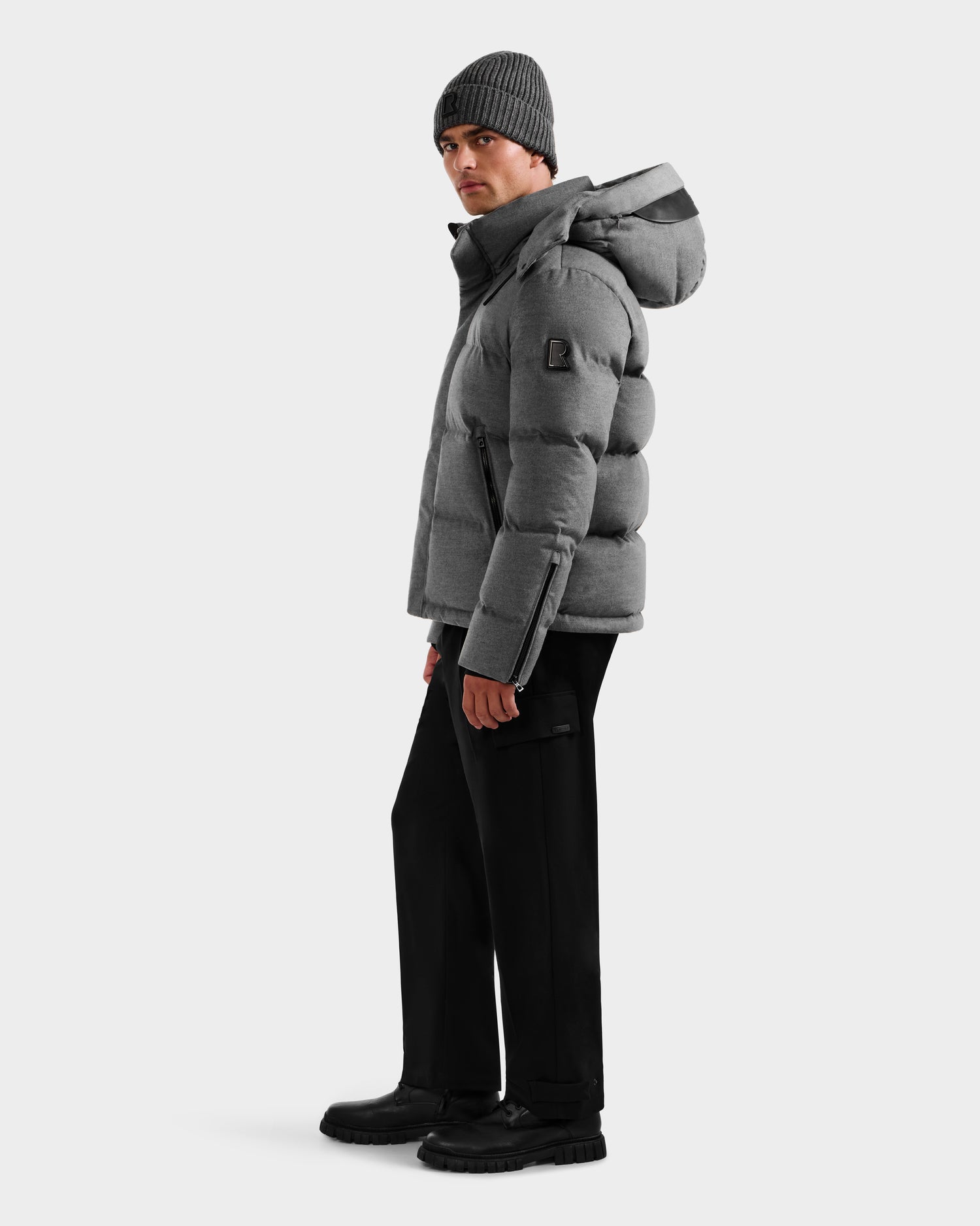 A person wearing the Rudsak JOEL W - 6125683 Heather Charcoal down jacket, paired with black pants, black boots, and a gray knit beanie, stands against a white background facing slightly to the side.