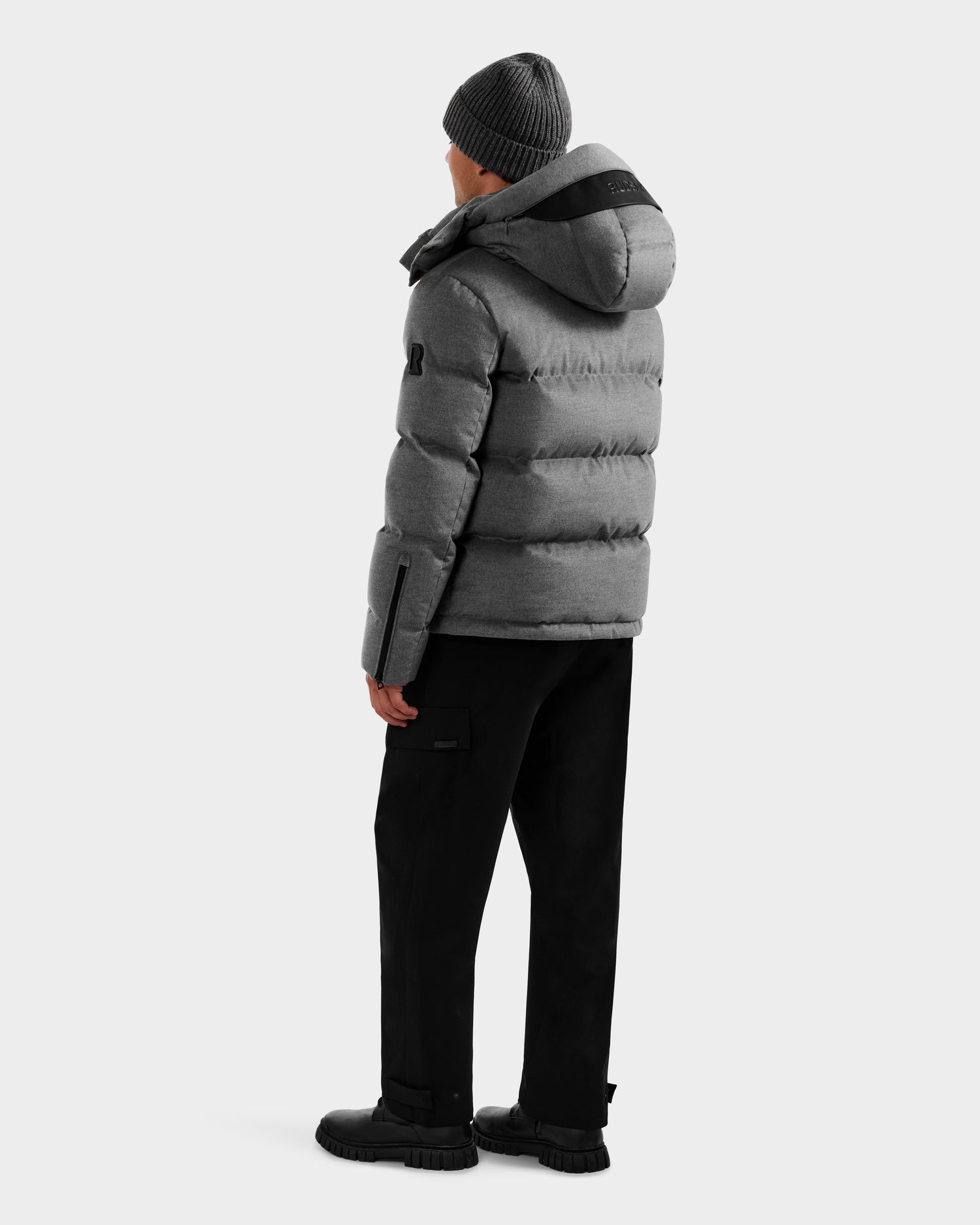 A person wearing the Rudsak JOEL W - 6125683 Heather Charcoal puffer, black pants, boots, and a gray beanie stands facing away against a plain white background.