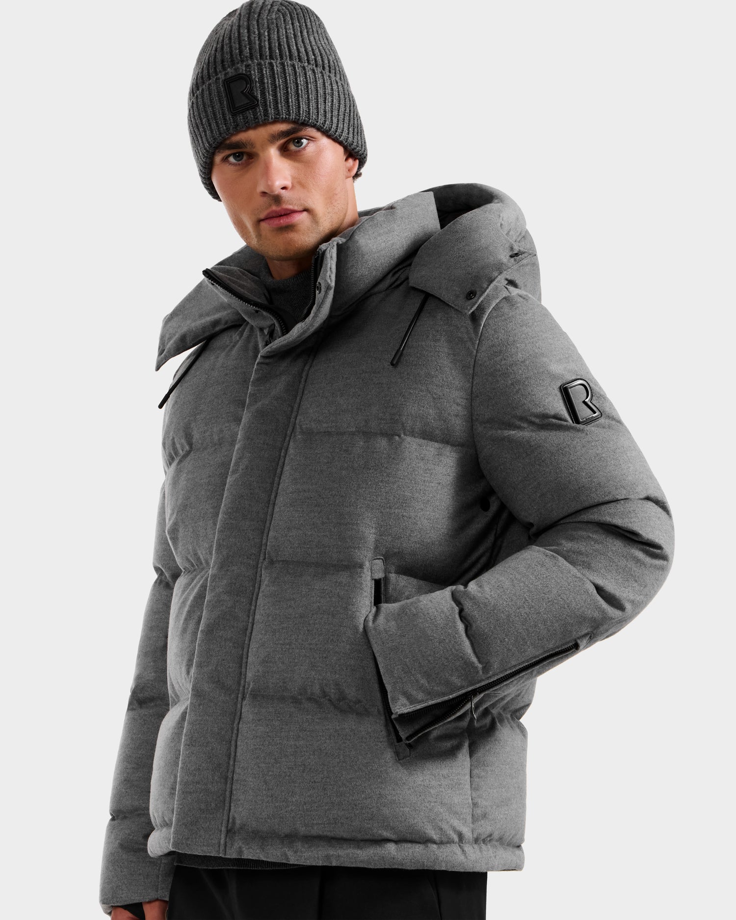 A person wears Rudsak’s JOEL W - 6125683 Heather Charcoal men’s down puffer with a removable hood and a matching gray knit beanie, both featuring the black R logo, standing against a plain light background.