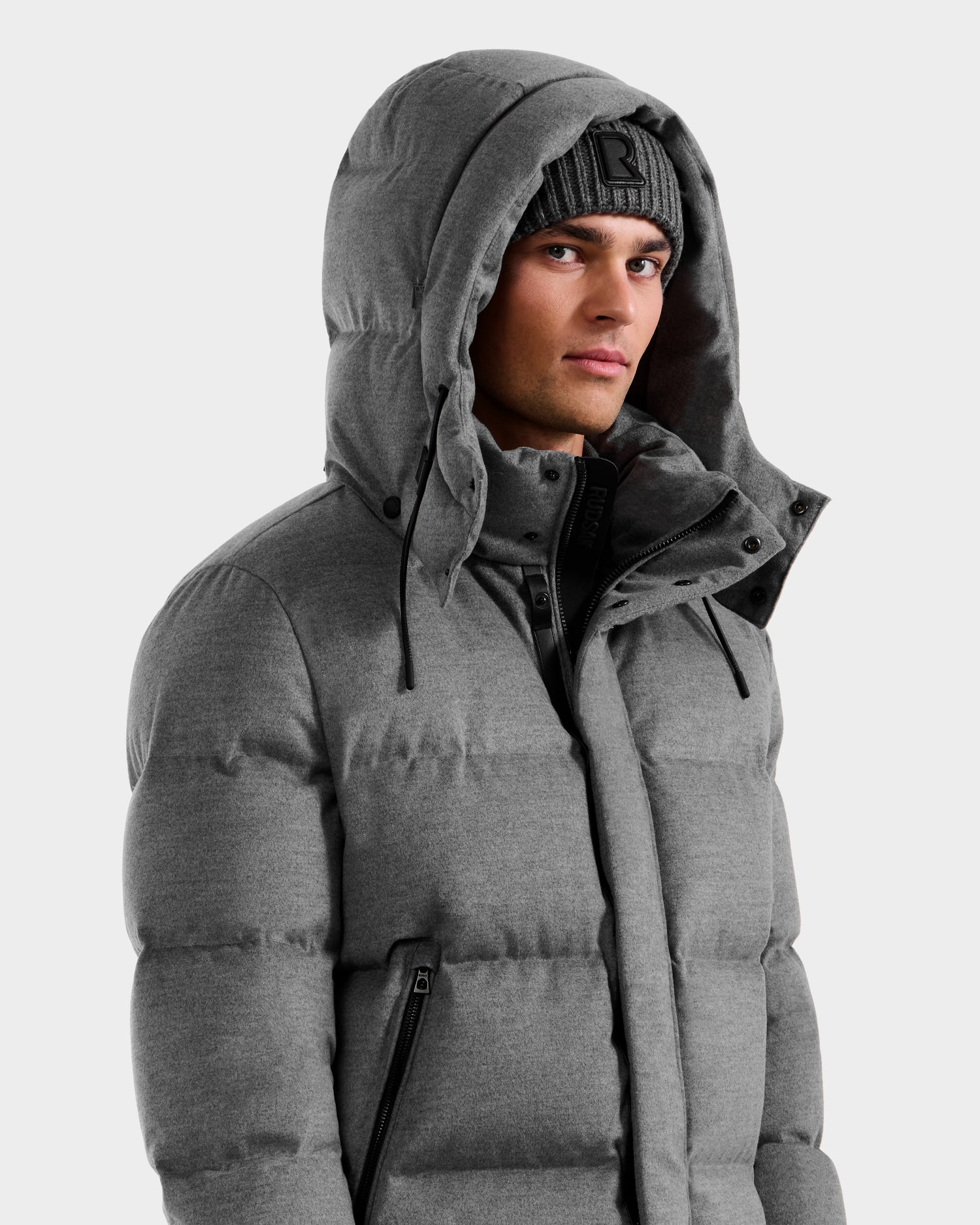 A man in the Rudsak JOEL W - 6125683 Heather Charcoal wool-blend down puffer and a matching gray knit beanie, hood up, gazes slightly to the side against a plain light background.