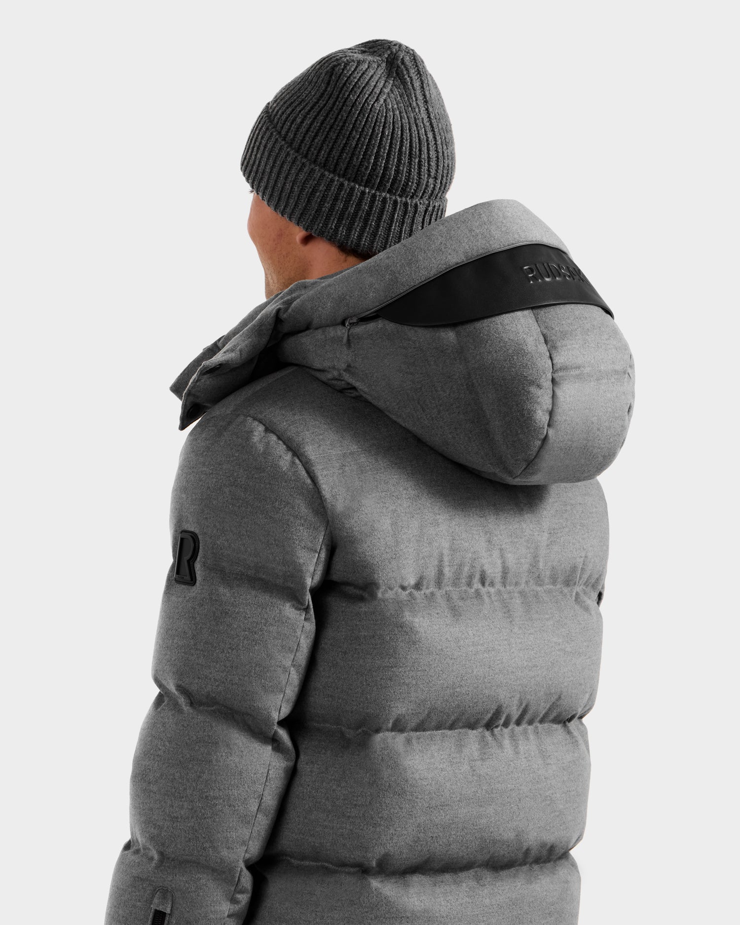 A person wearing the Rudsak JOEL W - 6125683 Heather Charcoal men’s wool-blend down puffer with removable hood and a gray knit beanie faces away from the camera against a plain light background.