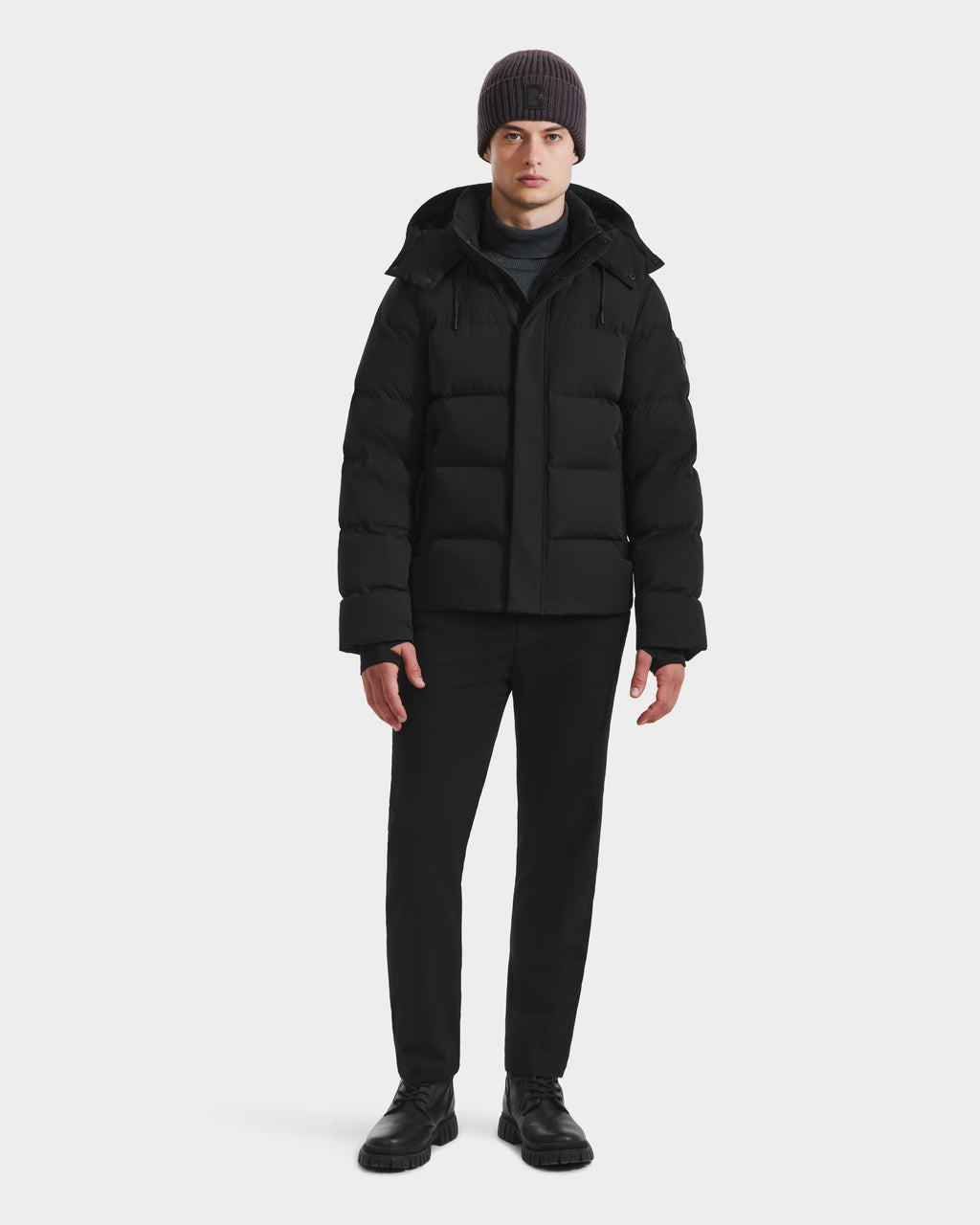 A person stands facing forward wearing the Rudsak JOEL - 6124605 Black puffer jacket with black pants, black shoes, and a dark knit beanie—showcasing stylish urban outerwear against a plain white background.