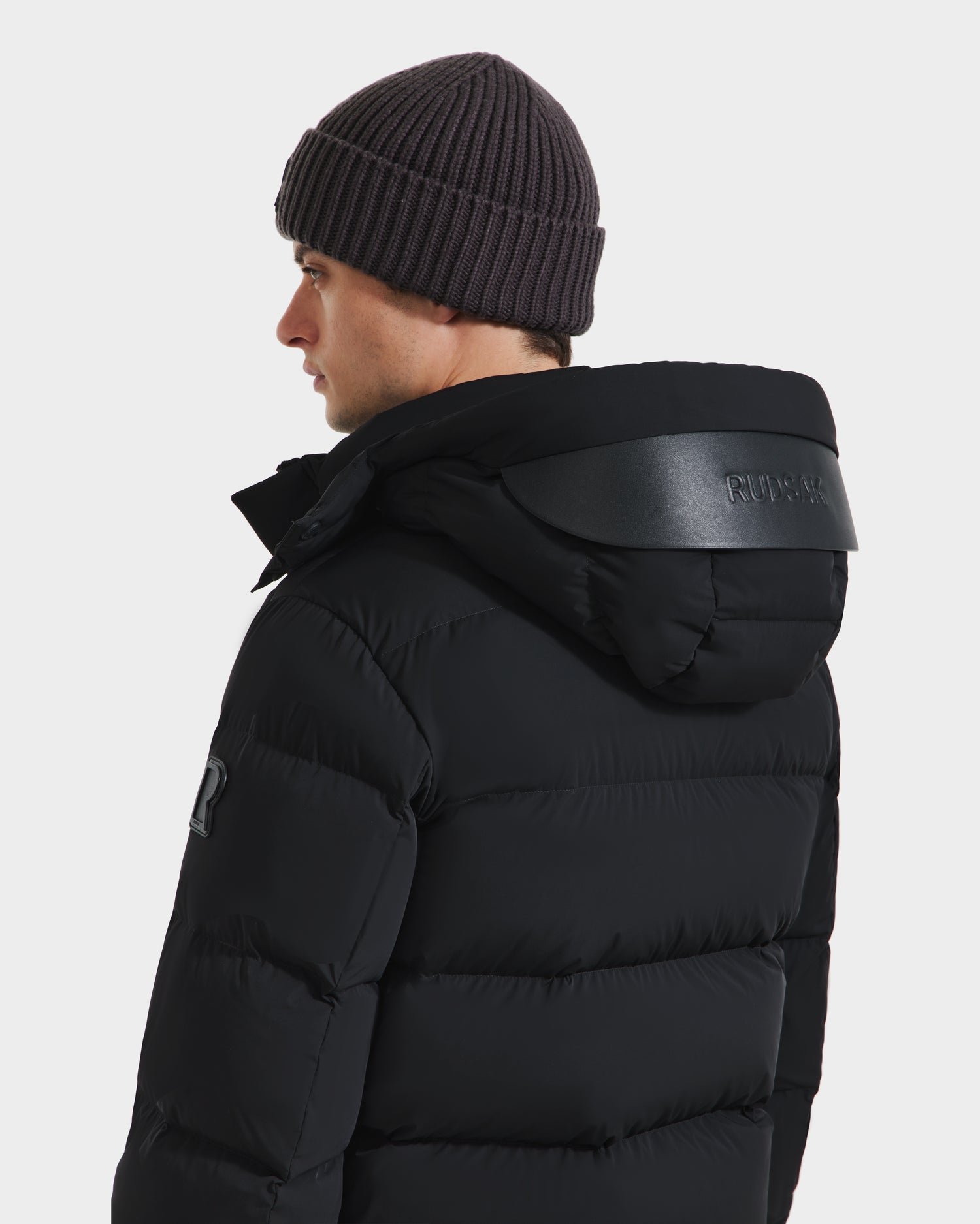 A person wearing the Rudsak JOEL - 6124605 Black men’s puffer jacket with a hood and a dark knit beanie, shown from the back and looking to the side against a plain white background.